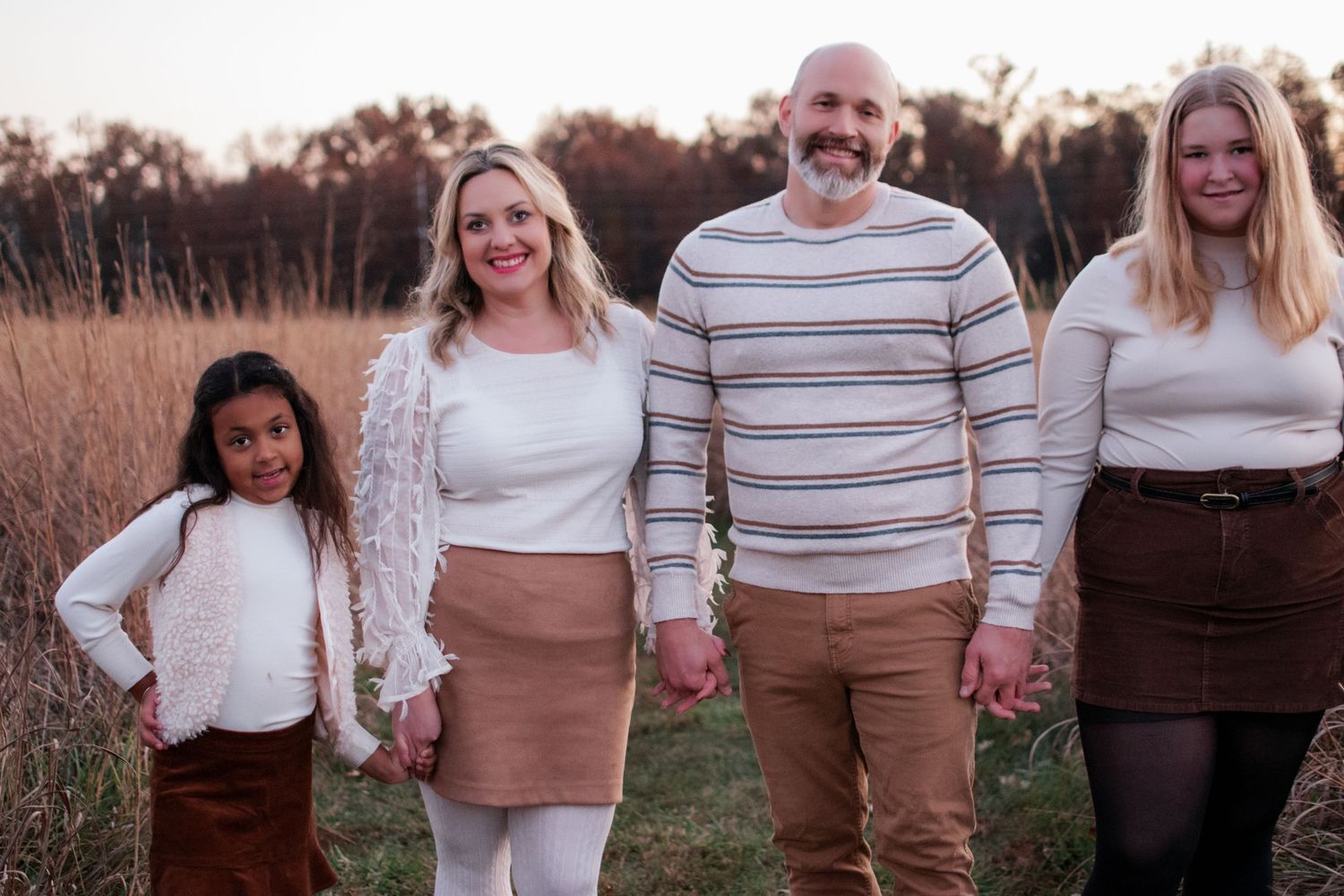 Happy family of four poses outdoors in autumn field, wearing coordinating neutral and brown outfits.