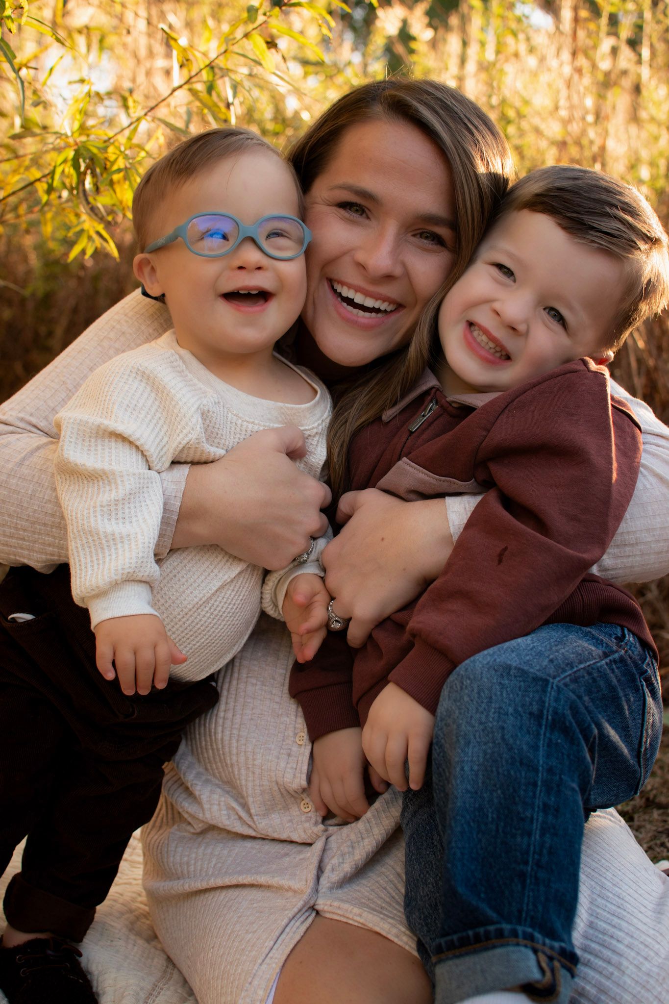 Happy mother hugging two laughing young sons outdoors in golden autumn light among fall foliage.