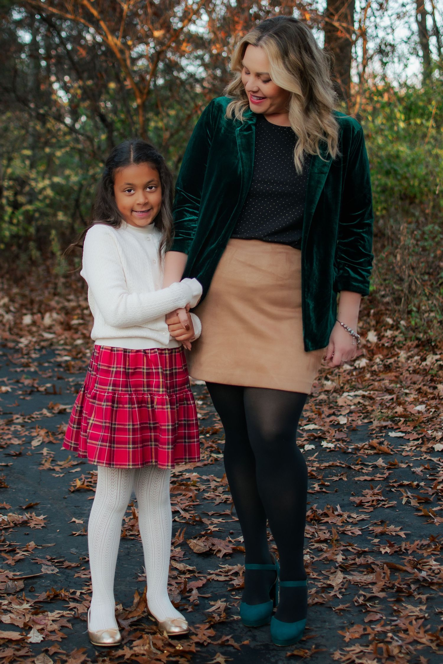 A woman in a green velvet blazer holds hands with a young girl in a red plaid skirt outdoors.