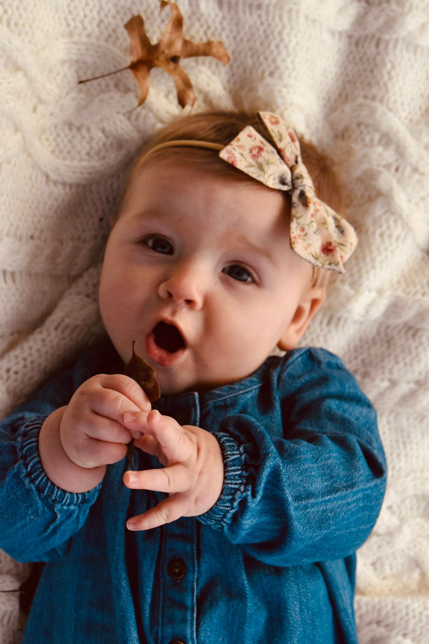 Baby girl in denim dress and floral bow headband lying on white knit blanket holding an autumn leaf.