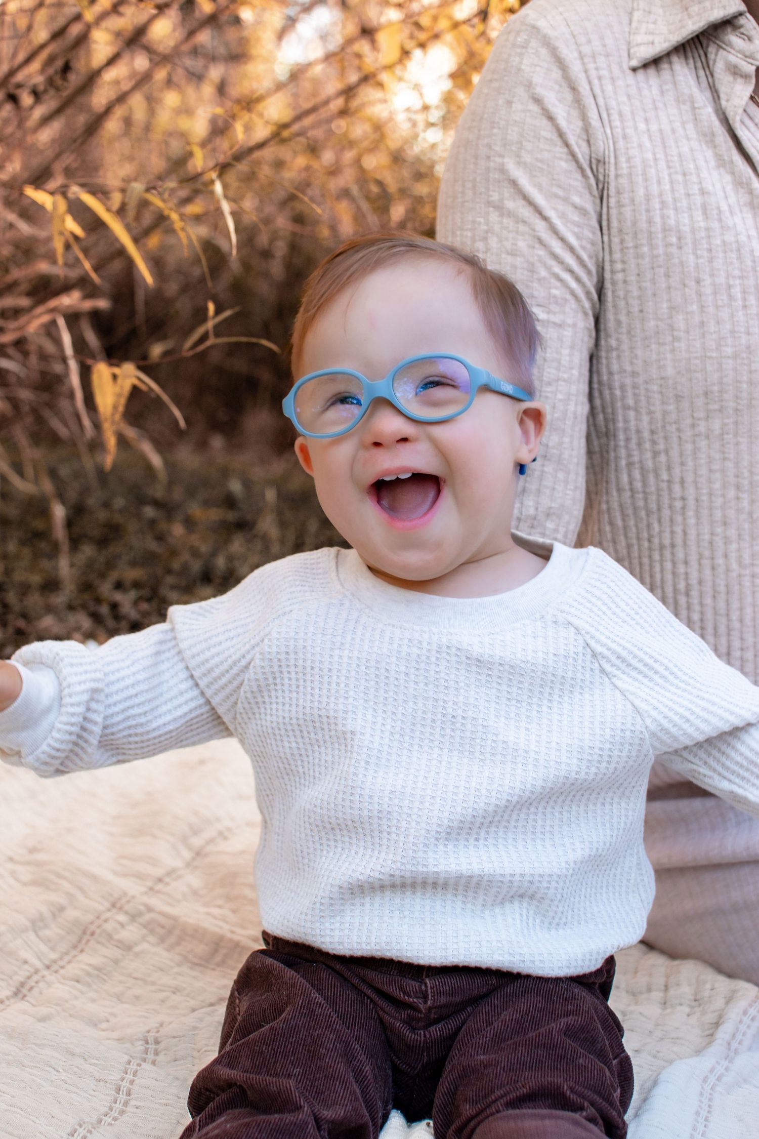 Joyful baby with blue glasses laughing outdoors in white sweater during golden autumn light.