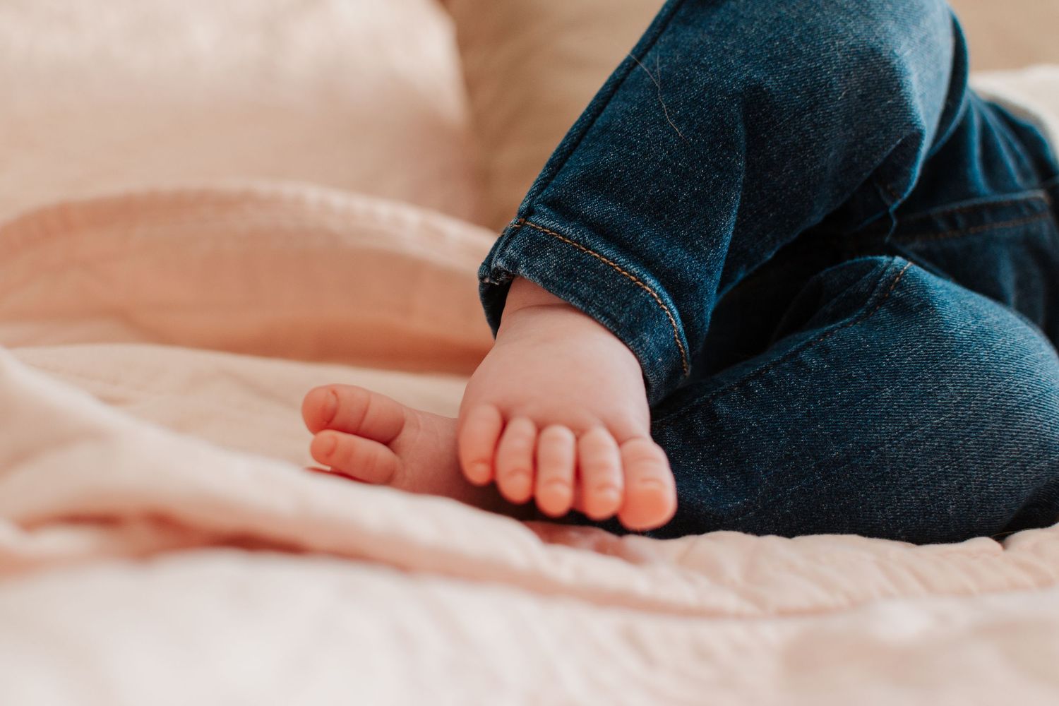 Close-up of a baby's tiny bare feet in rolled-up denim jeans on a pink blanket.