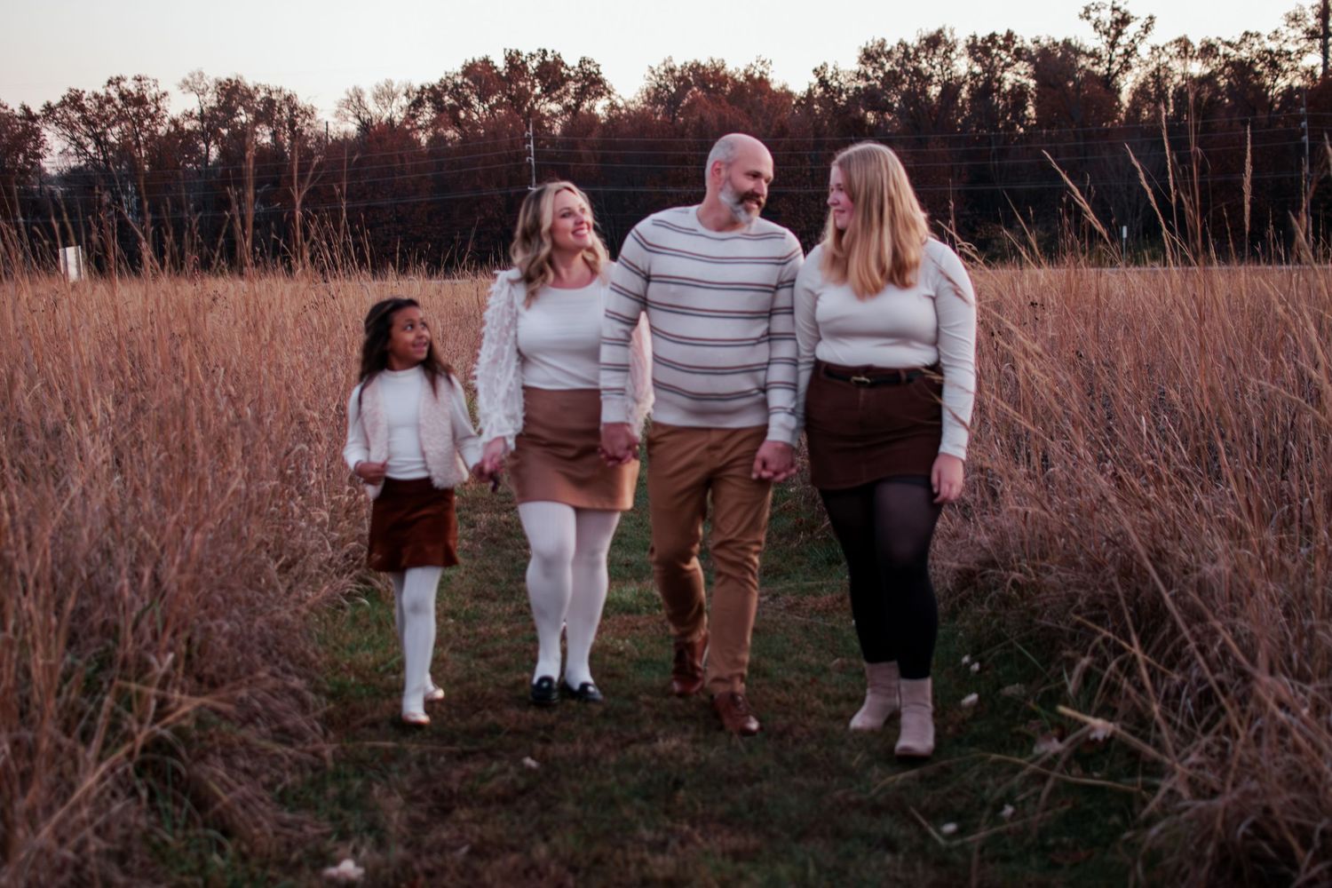 A cheerful family of four walks hand-in-hand through a golden autumn field at sunset.