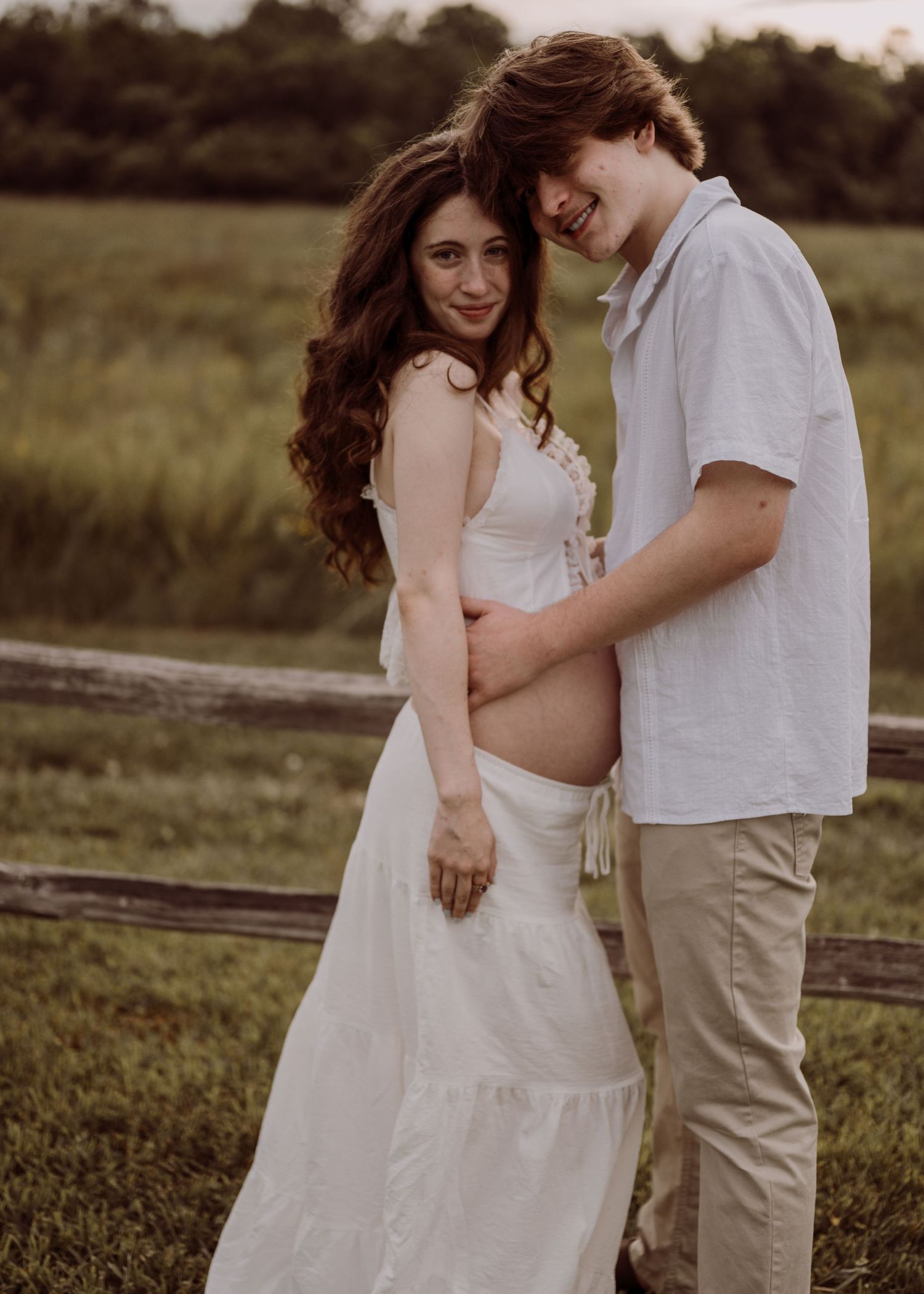 Couple in white outfits posing for maternity photo in green field, woman in white dress showing baby bump.