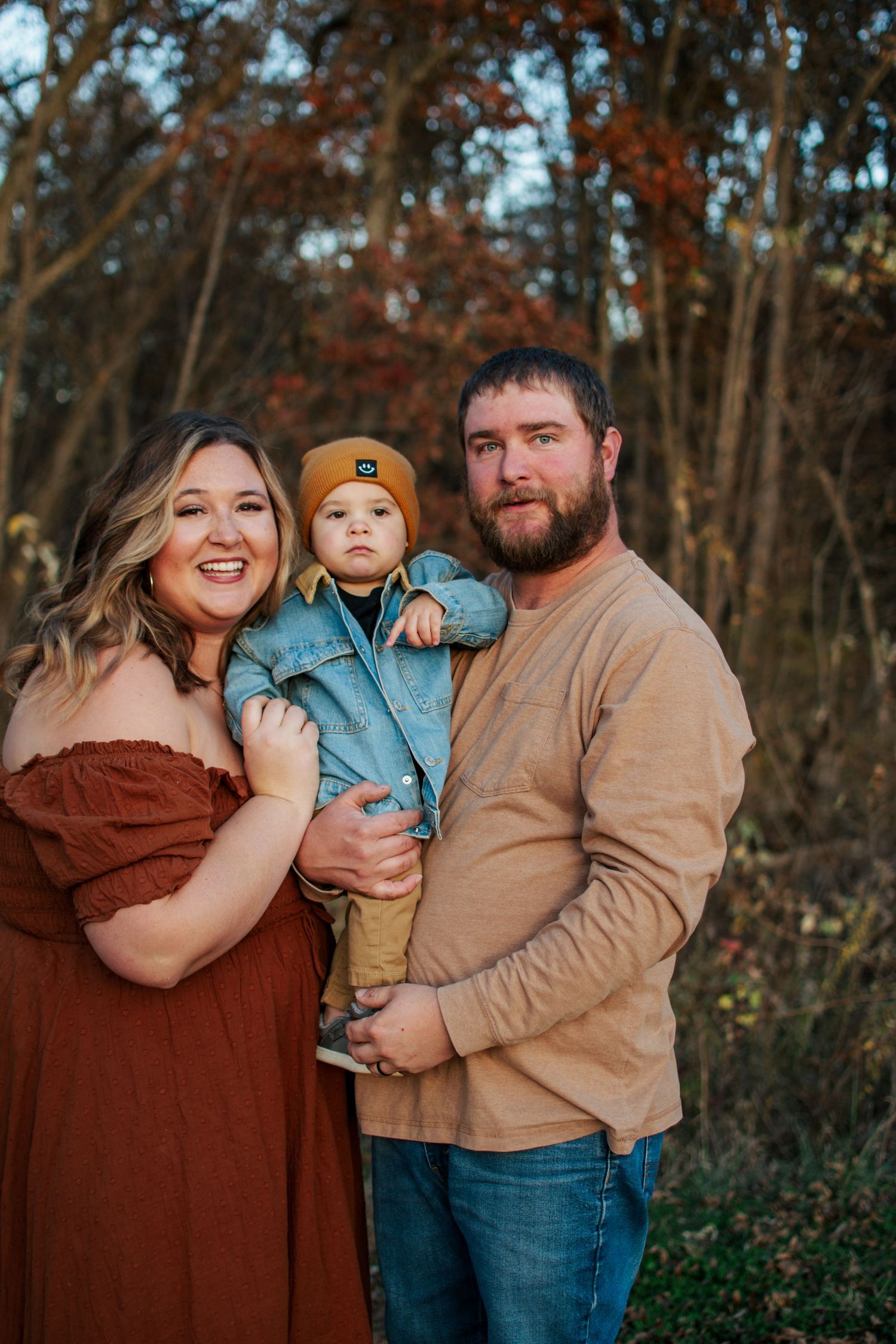 Young family of three posing outdoors in fall foliage, baby wearing denim jacket and mustard hat.