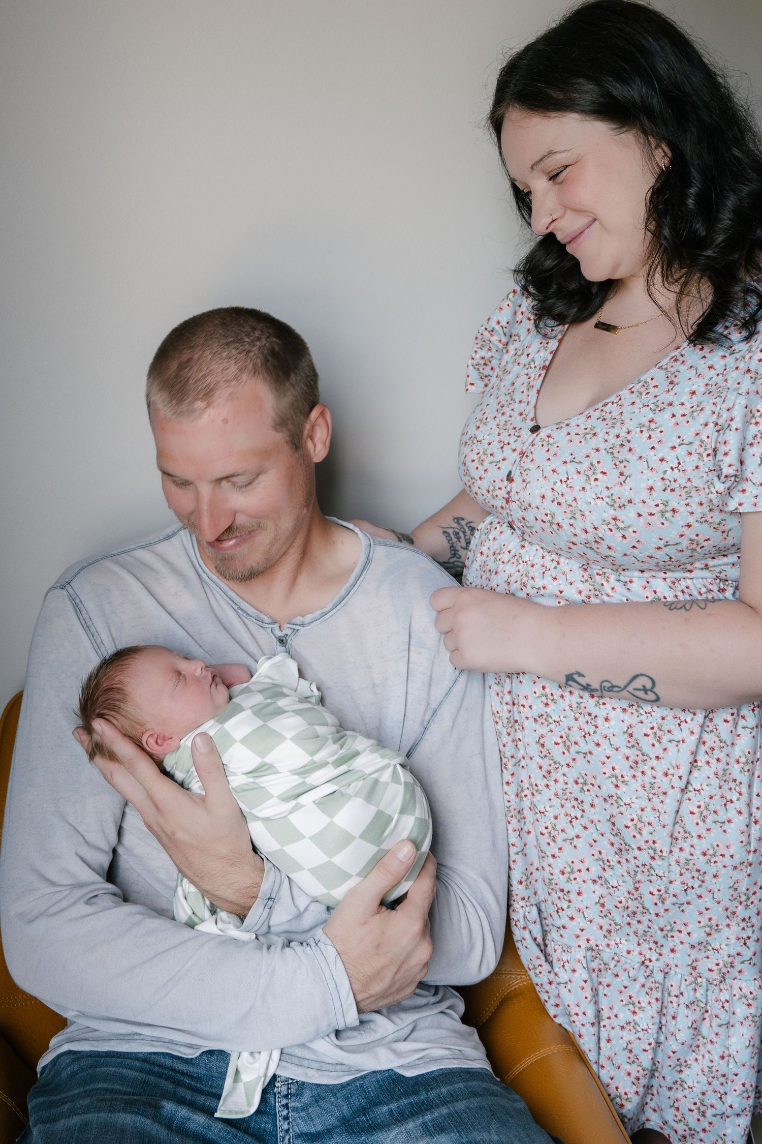 New parents smiling lovingly at their swaddled newborn baby, father seated holding infant, mother standing beside them.