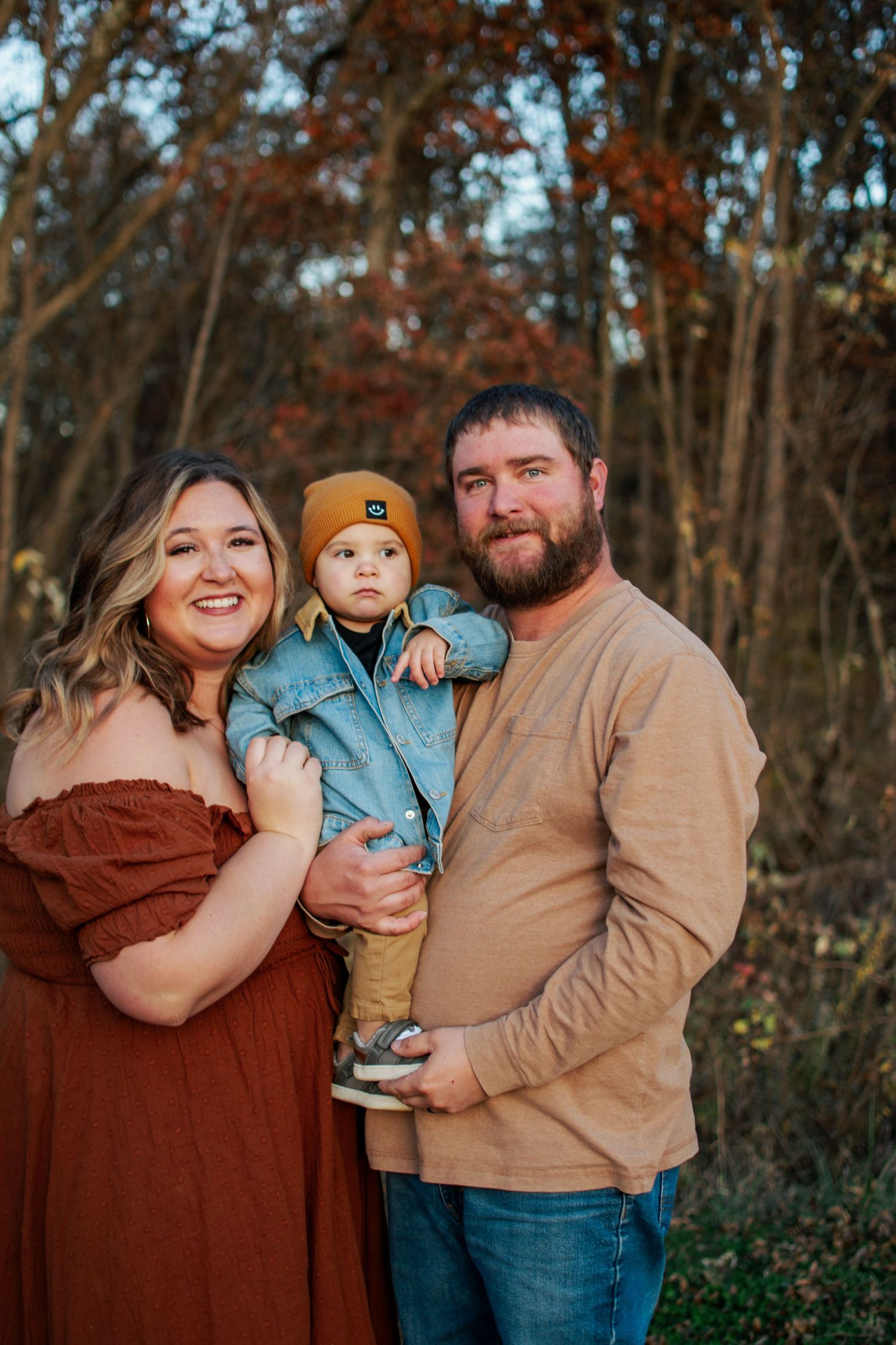 Cheerful family of three posing outdoors in fall foliage, mom in rust dress, dad holding toddler in denim jacket.