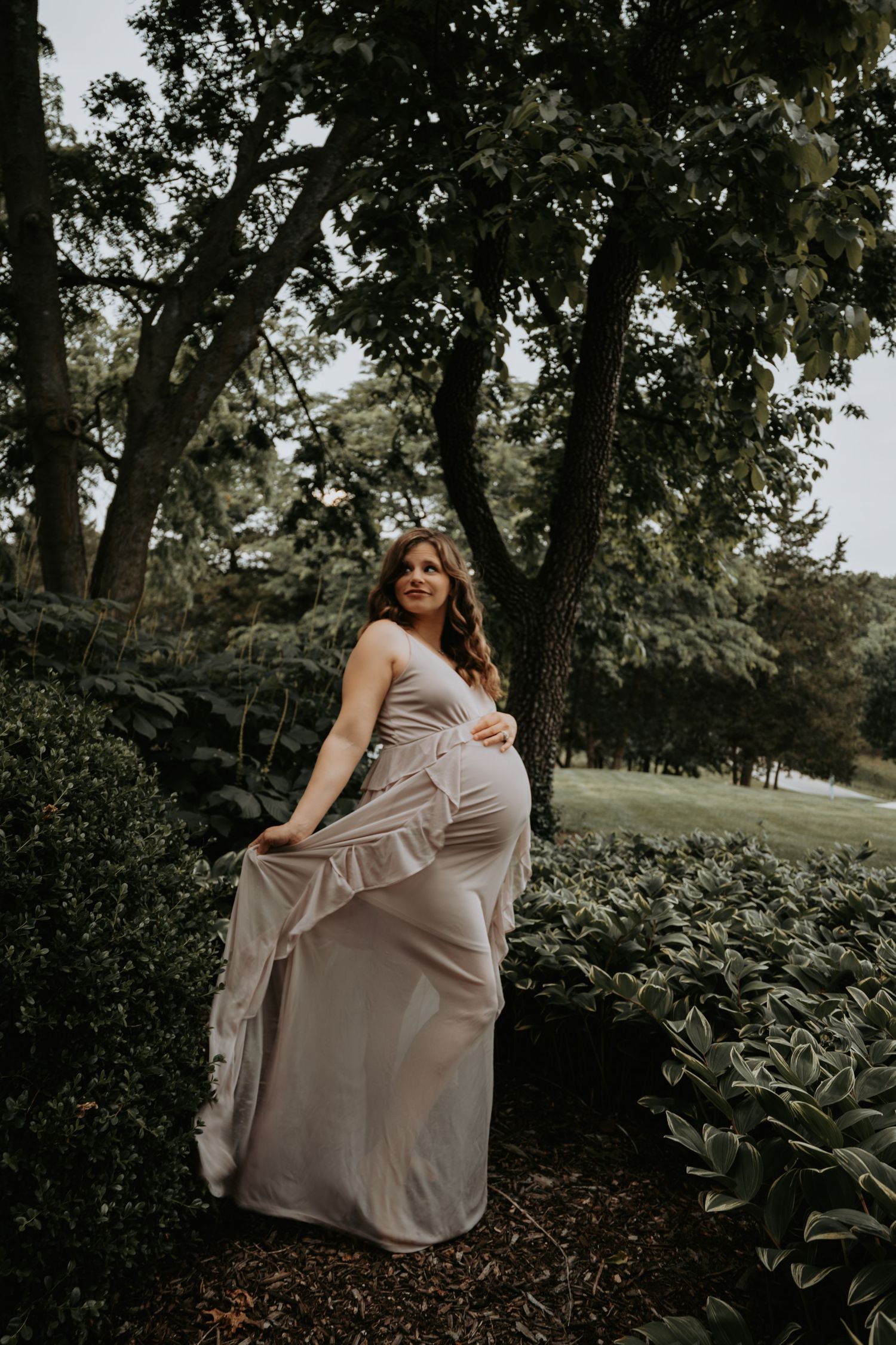 Smiling pregnant woman in lavender gown posing beside lush garden shrubs under large trees.
