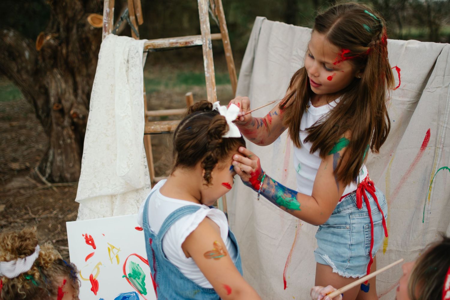 A young girl with paint on her face and arms places a bow on a woman's hair outdoors near canvas backdrops.