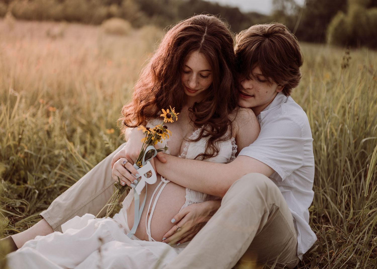 Couple sitting in golden meadow holding baby shoes and sunflowers during maternity photo session.