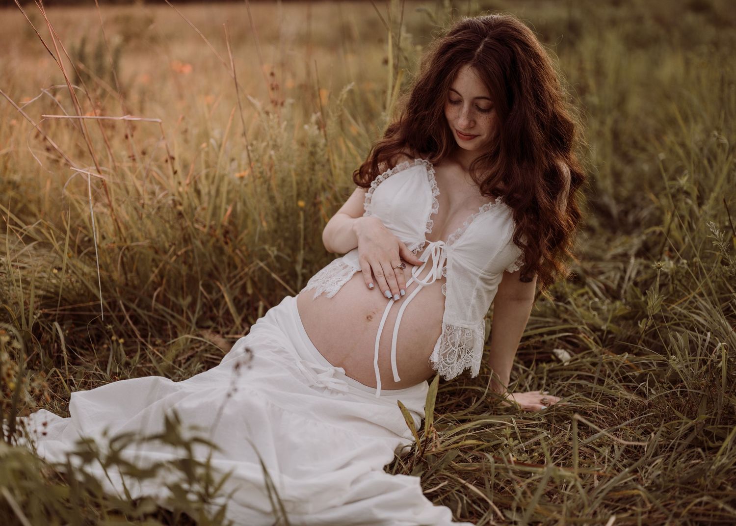 A pregnant woman in a white lace dress sits gracefully in a sunlit meadow cradling her baby bump.