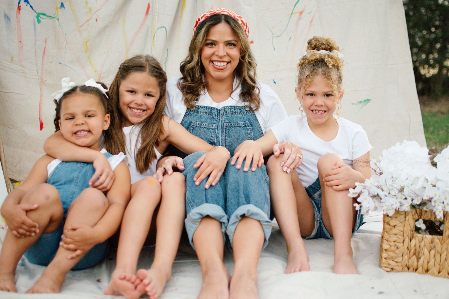 A smiling woman and three young girls sit together barefoot in front of a colorful painted teepee backdrop.
