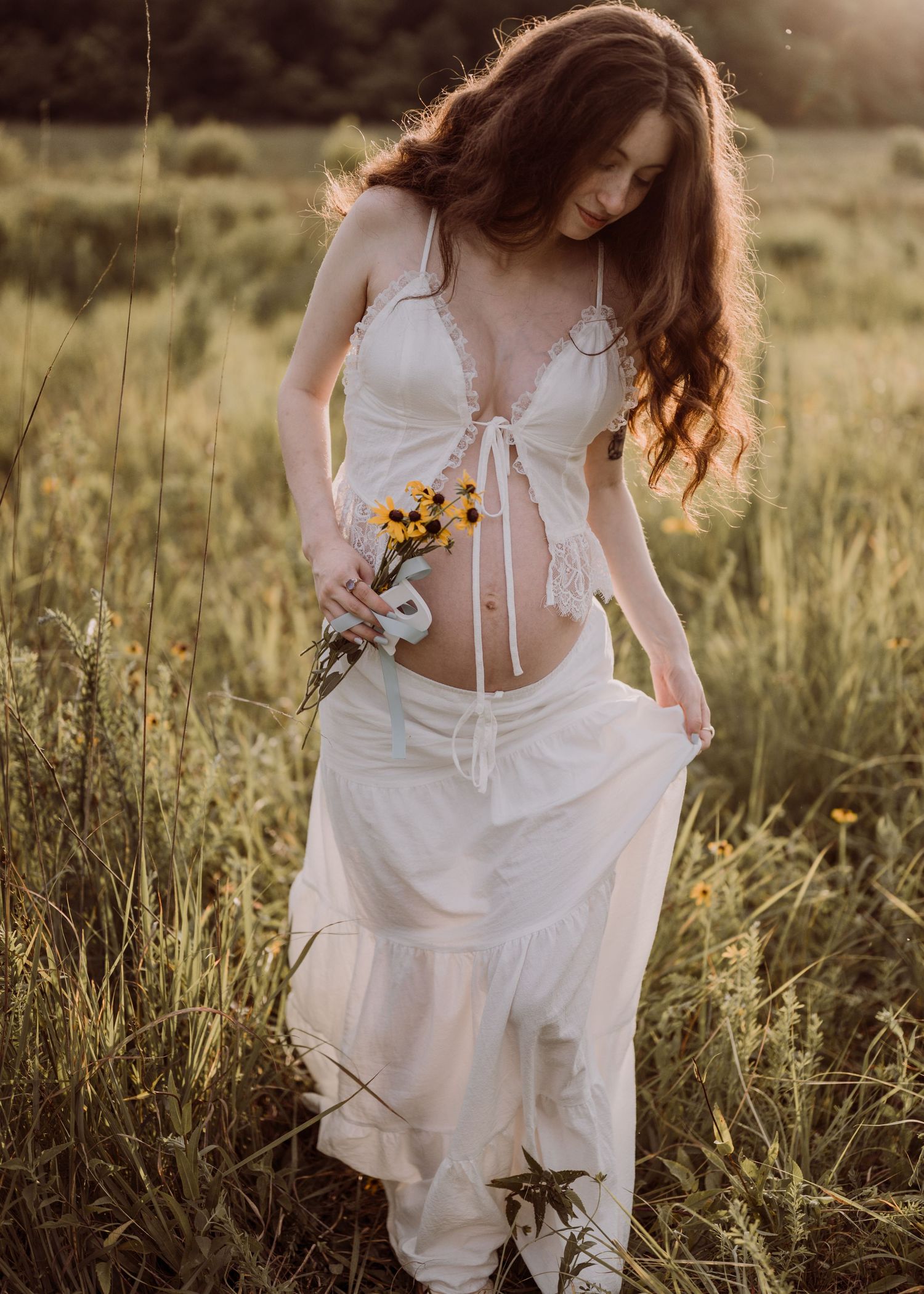 Pregnant woman in white boho dress holding wildflowers in a sunlit meadow field during golden hour.