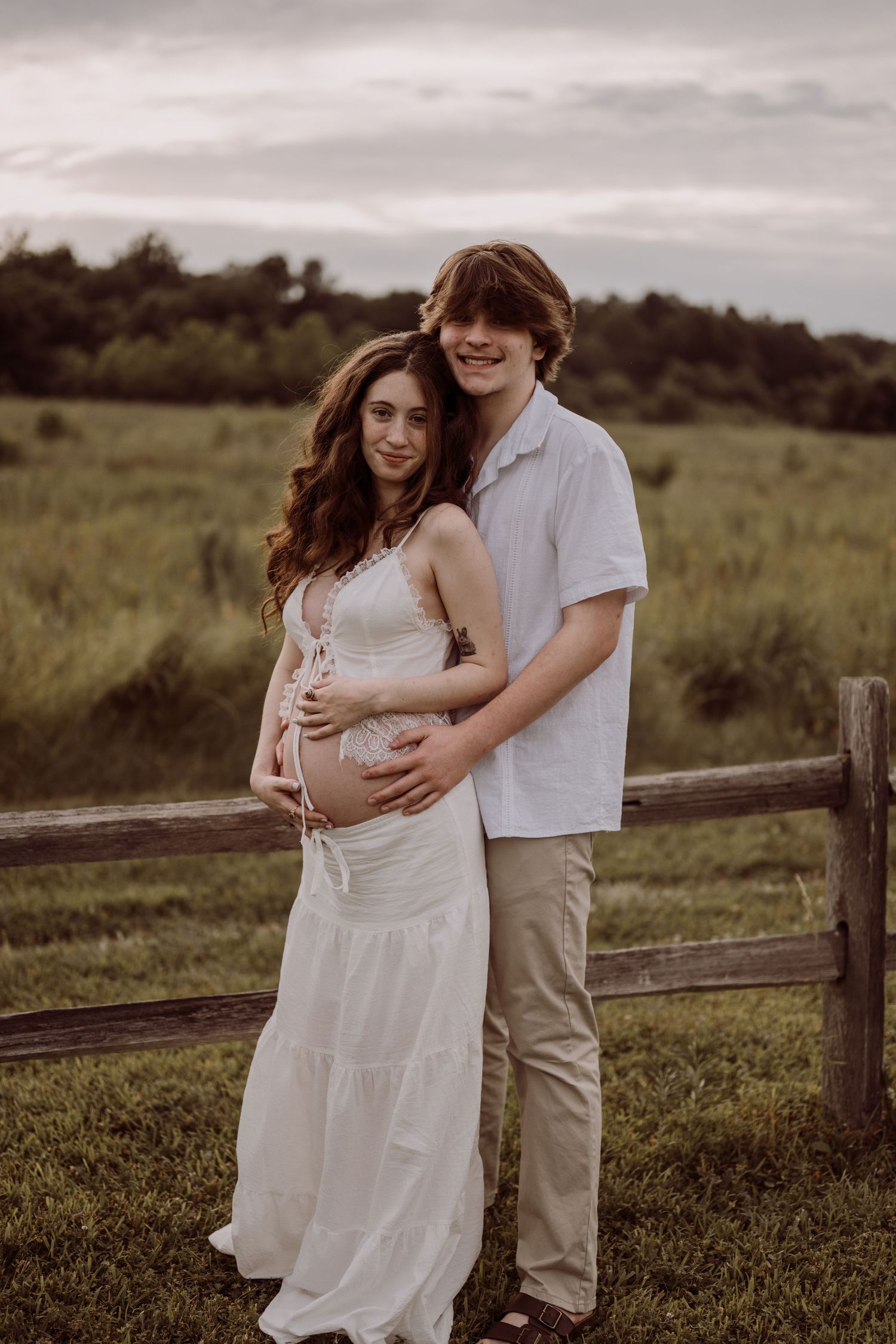 Young couple posing outdoors in a field, woman in white dress showing baby bump, man in white shirt embracing her.