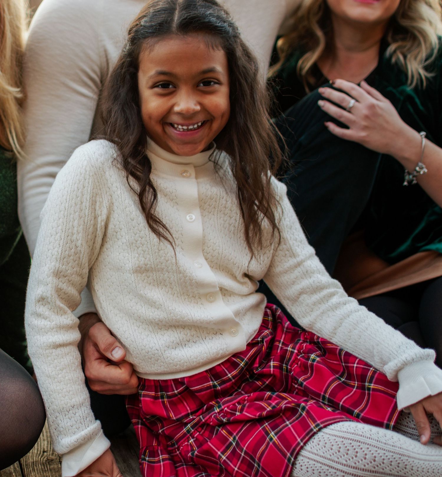 A smiling young girl in a cream sweater and red plaid skirt sits happily during a family photo session.