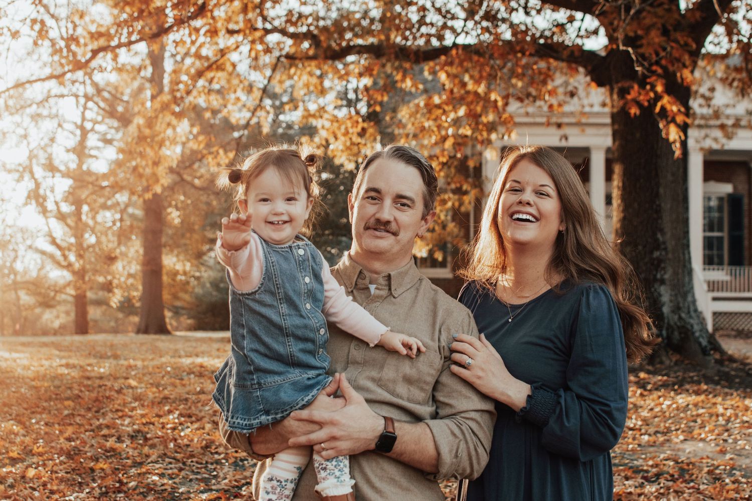 Happy family of three posing outdoors among autumn leaves with a house in the background.