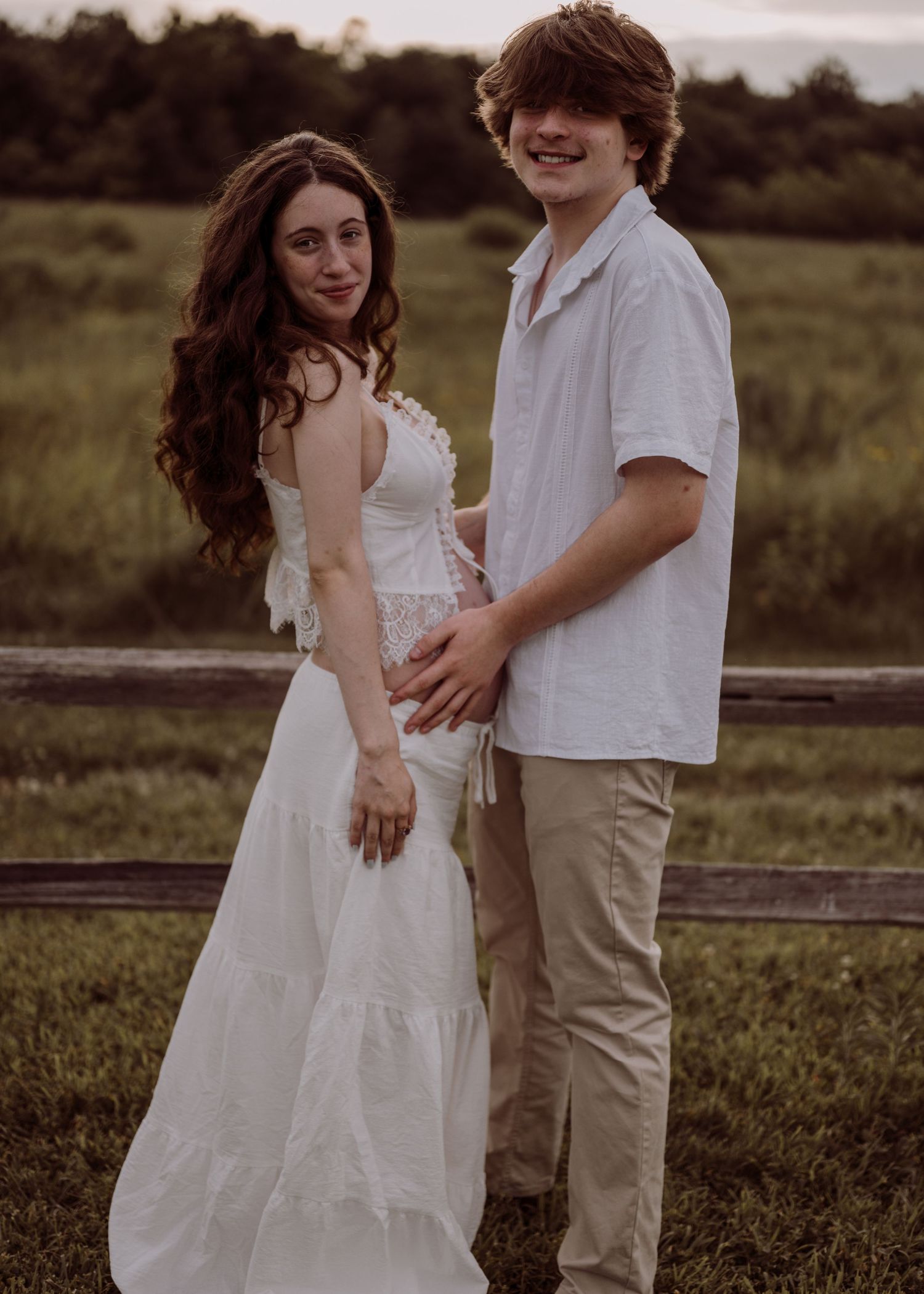 Young couple holding hands outdoors near wooden fence, woman in white dress and man in white shirt smiling.