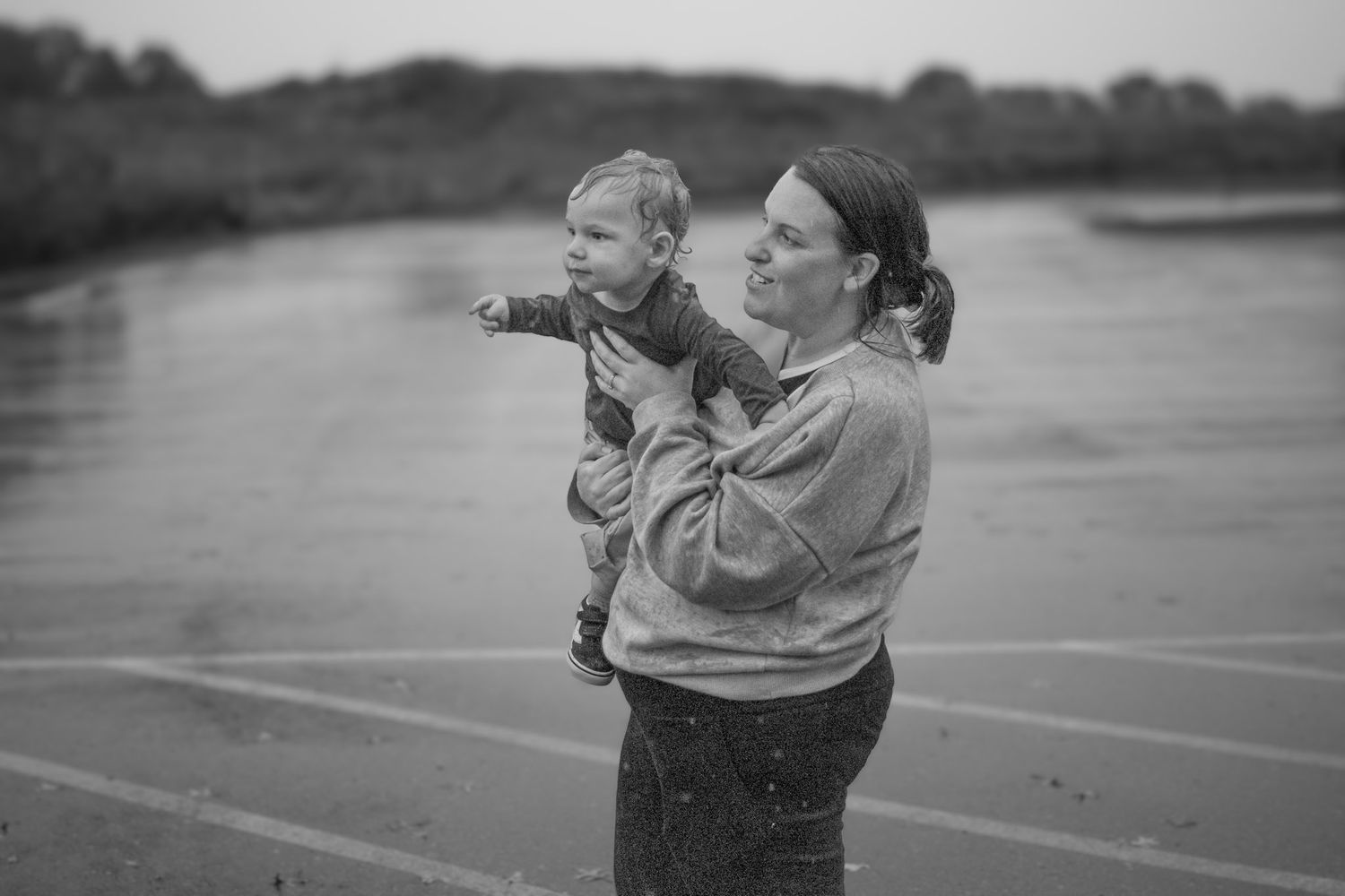 Black and white photo of mother holding baby in the rain near a reflective wet surface outdoors.