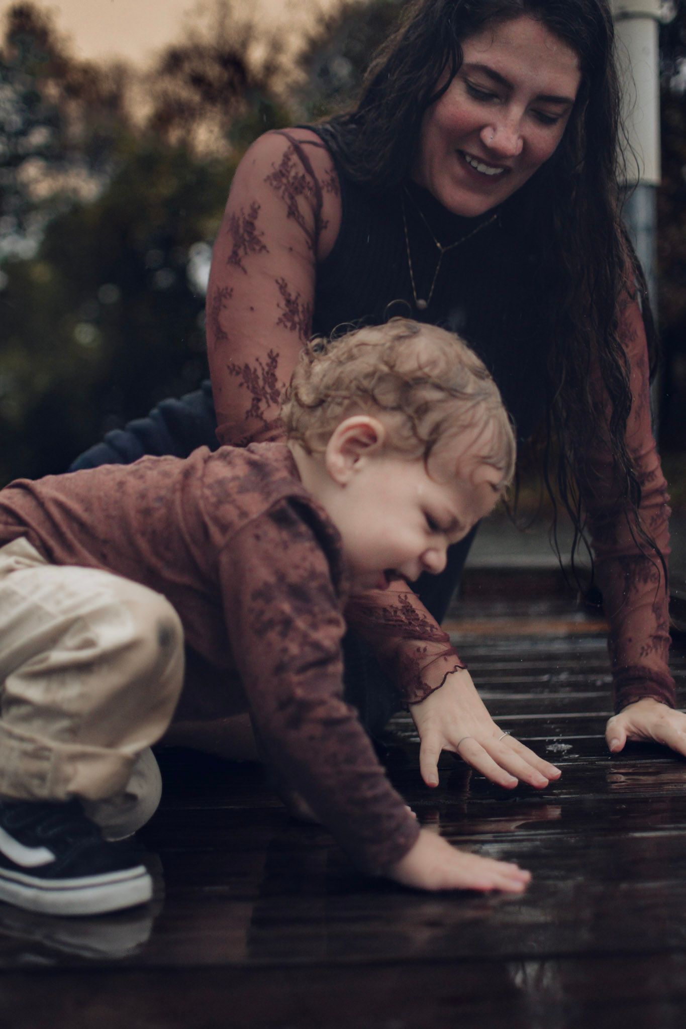 Smiling mother watching curious toddler boy touch wet wooden deck surface outdoors in autumn.