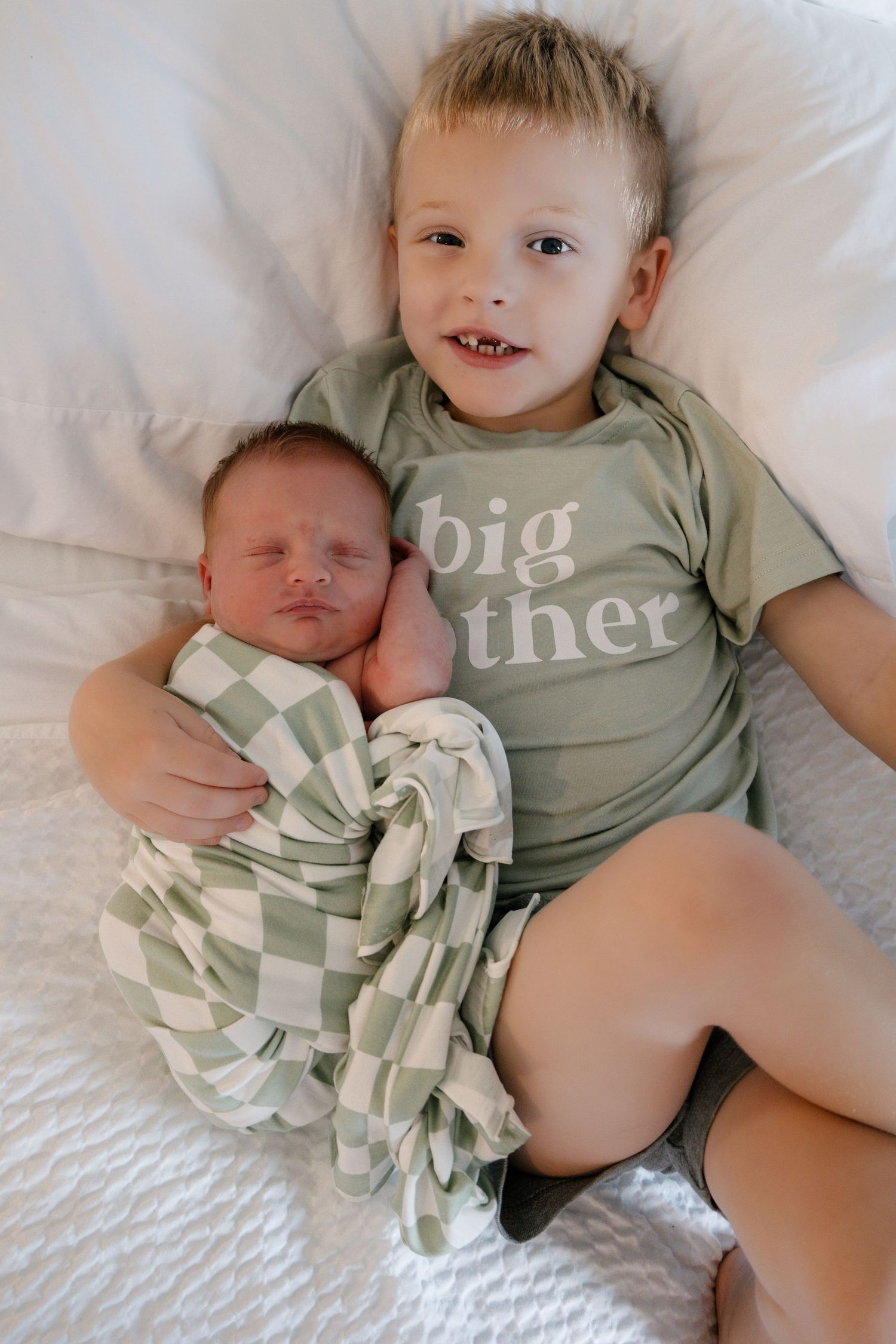 Young boy wearing a 'big brother' green shirt lying on white bed holding a swaddled newborn baby sibling.