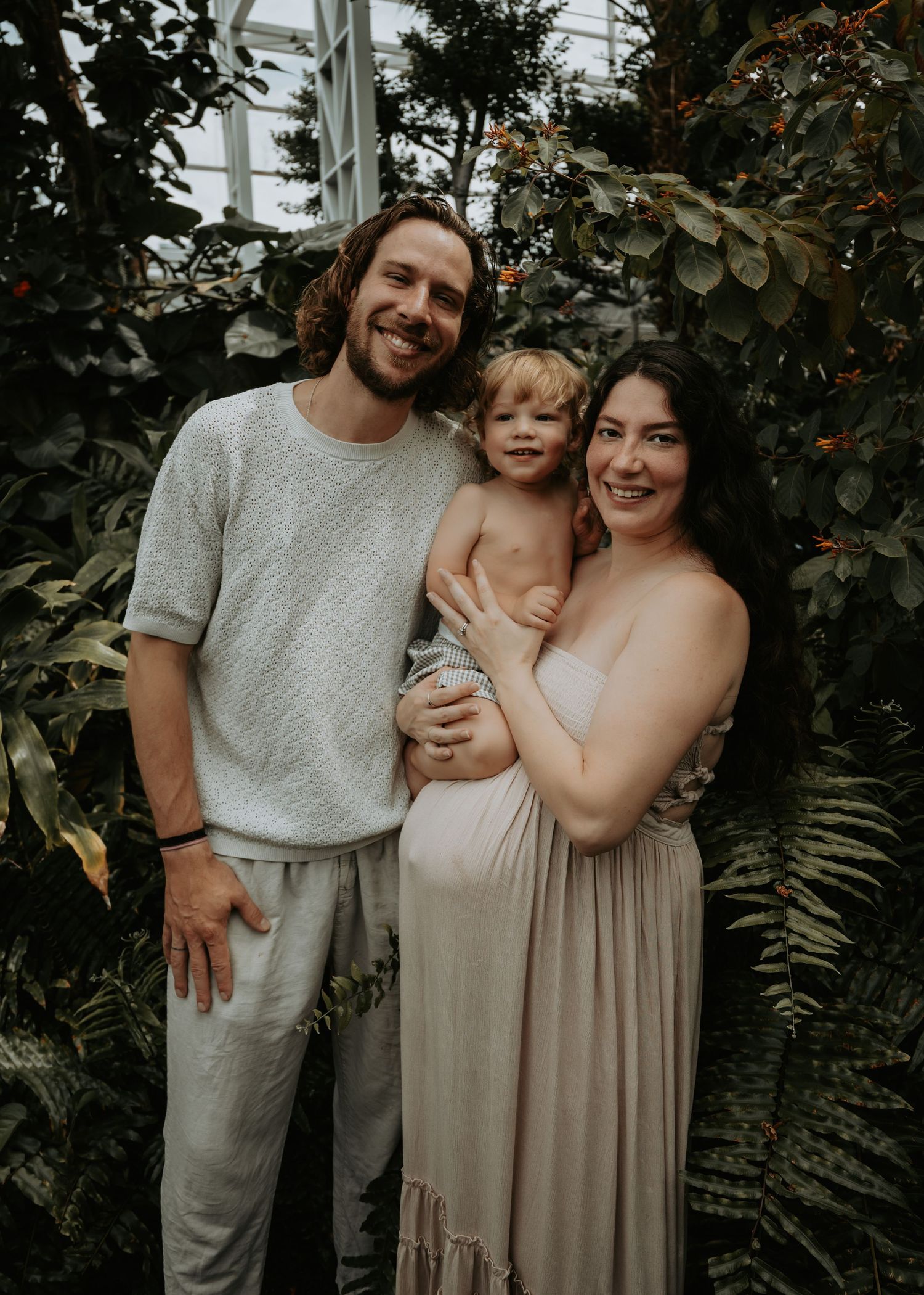 A smiling family of three poses among lush greenery, with parents holding a toddler in a greenhouse setting.