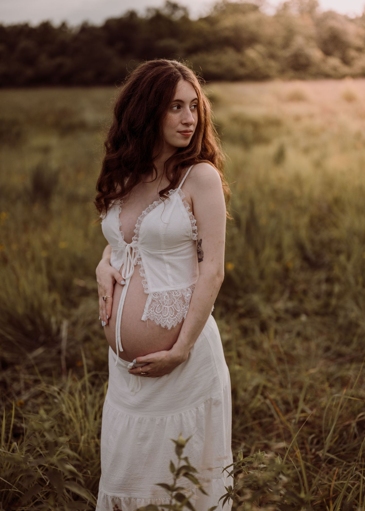 A pregnant woman in a white lace maternity gown cradles her baby bump in a golden sunset field portrait.