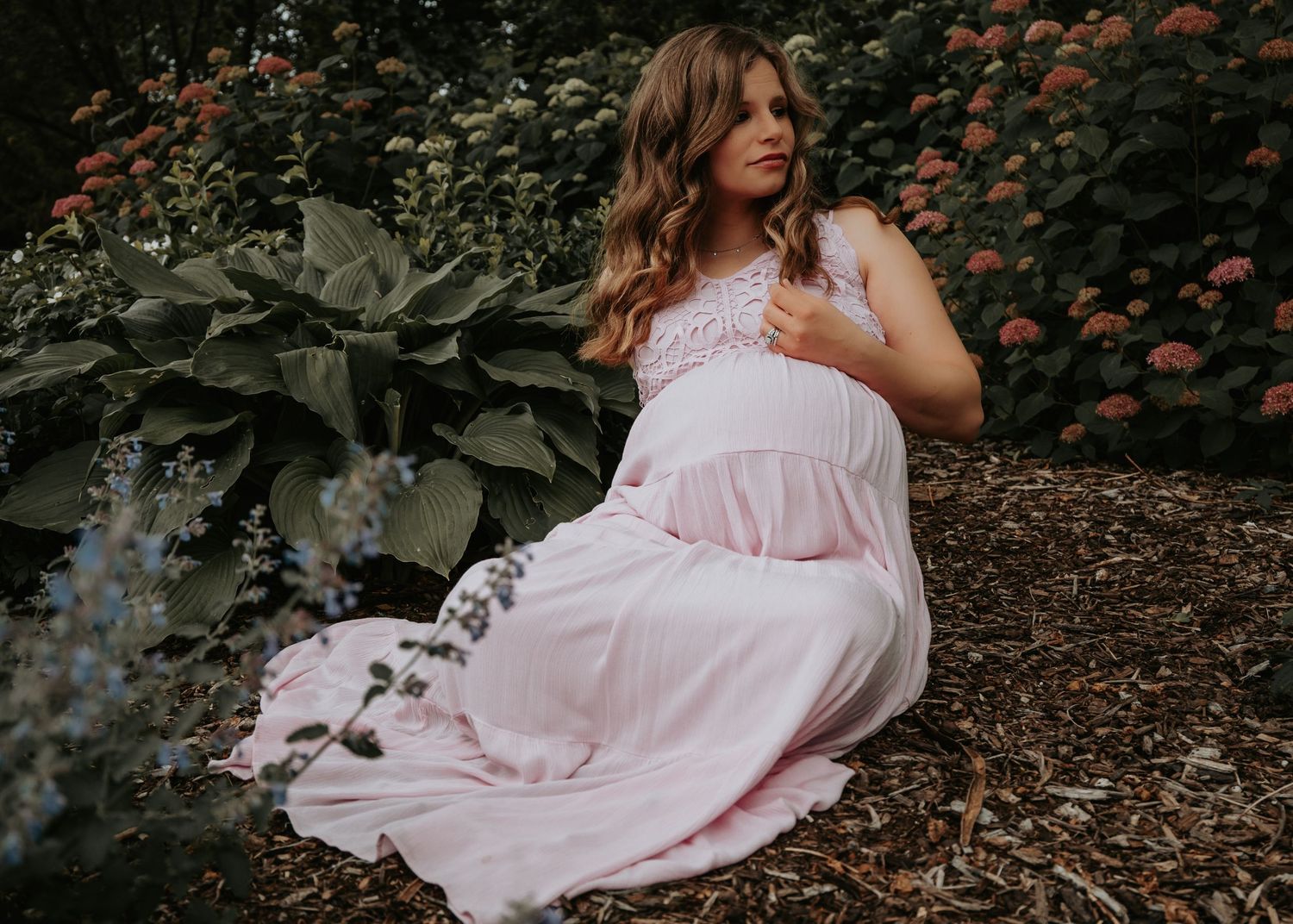 Pregnant woman in blush pink lace maternity gown sitting peacefully in a garden setting.