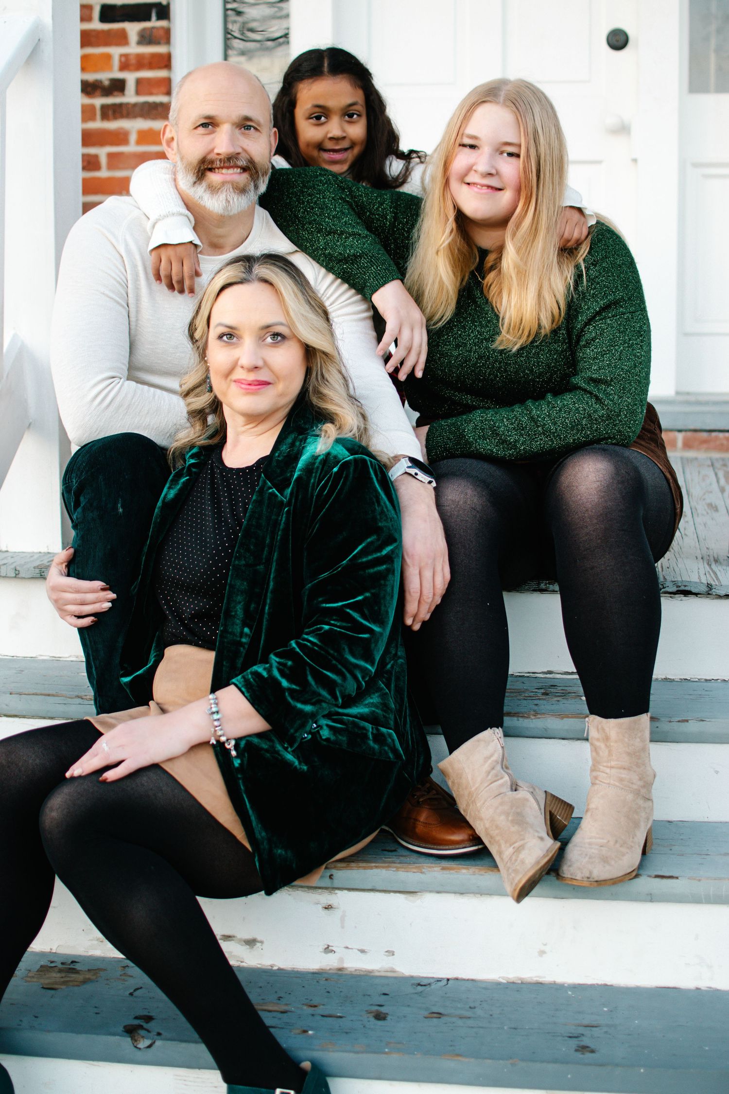A family of four sits together on white porch steps wearing coordinated green and neutral holiday outfits.