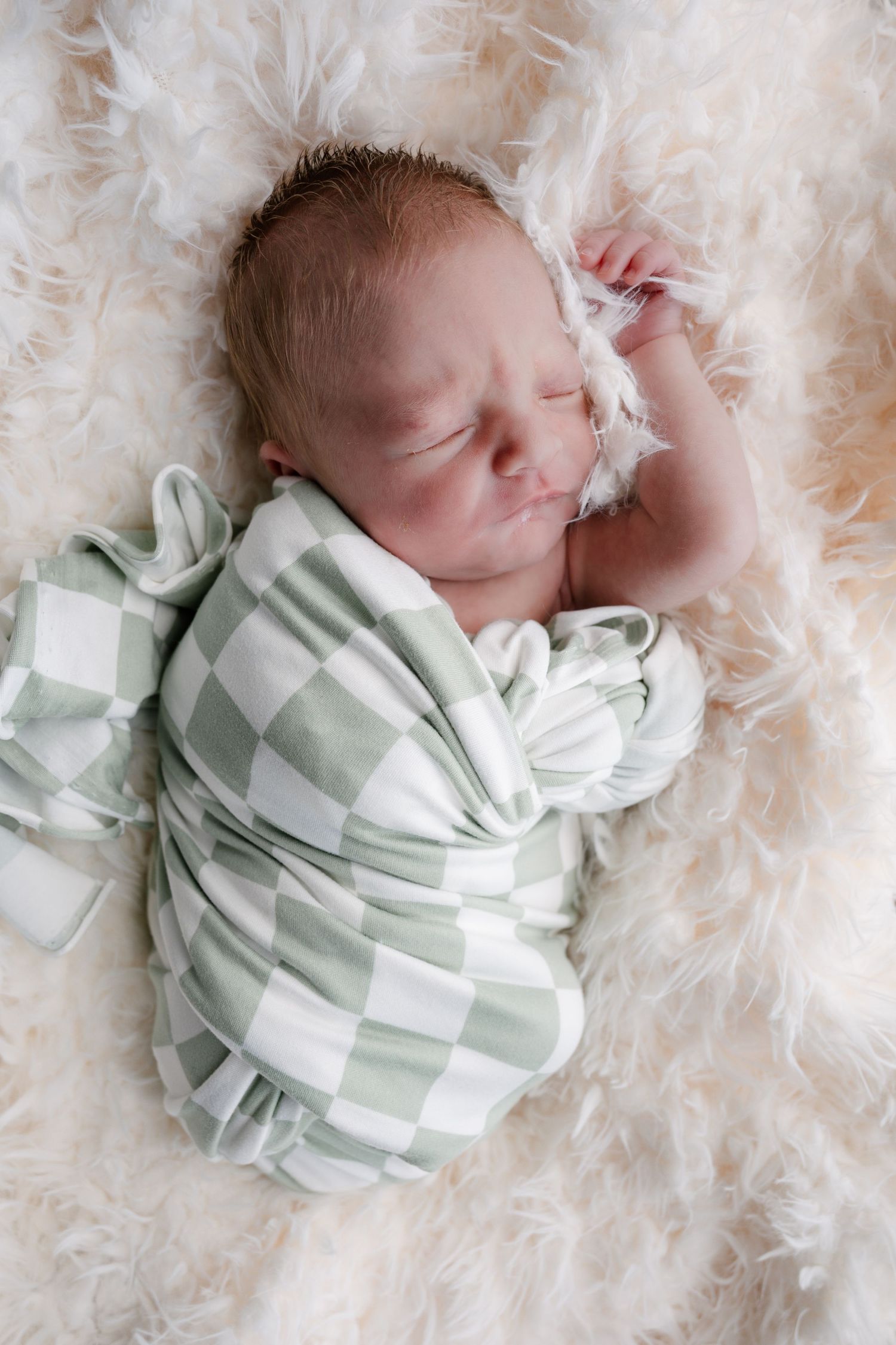 Sleeping newborn baby swaddled in green and white checkered blanket resting on fluffy white fur.