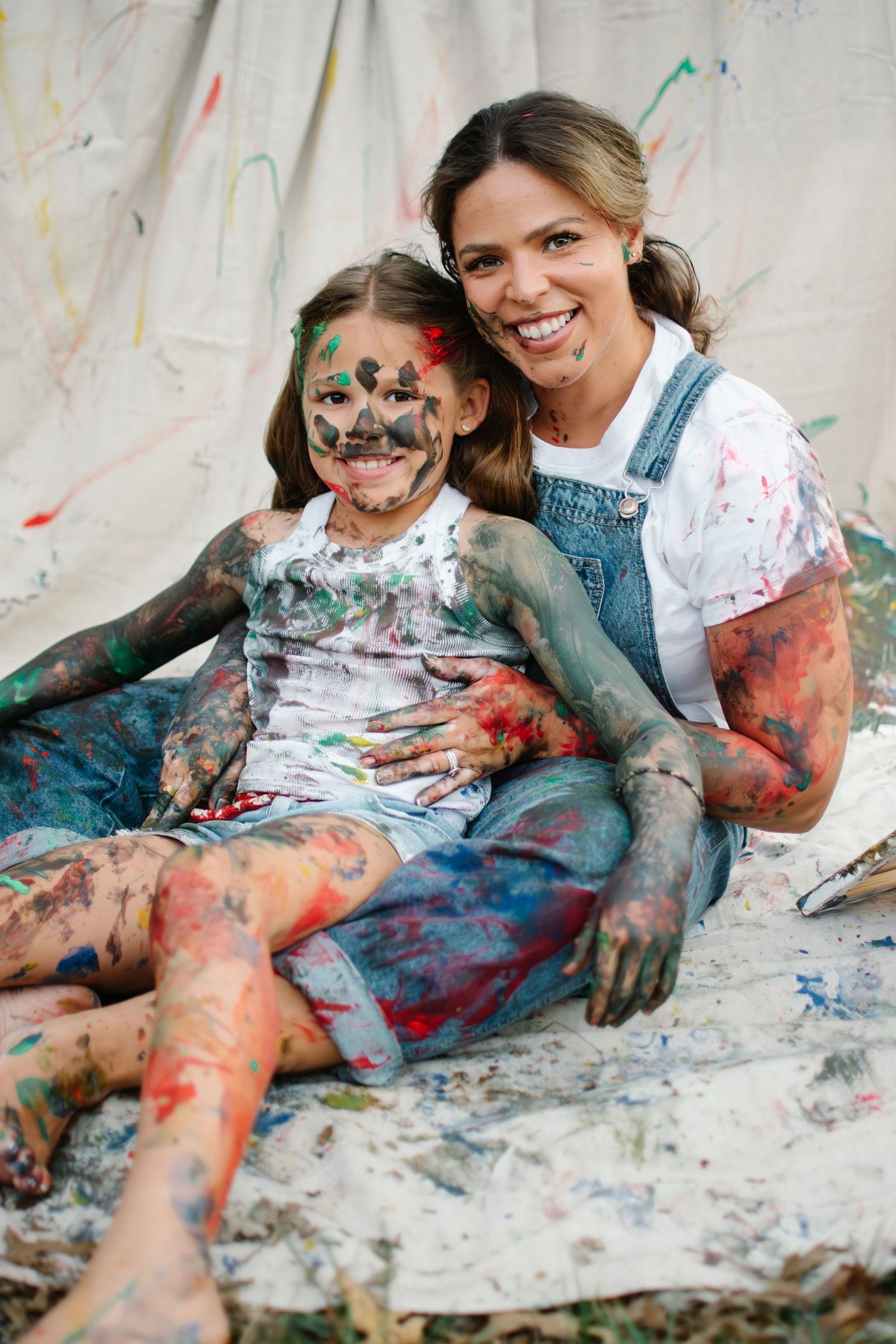 A smiling woman and paint-covered girl sit together on a splattered canvas, both laughing and covered in colorful paint.