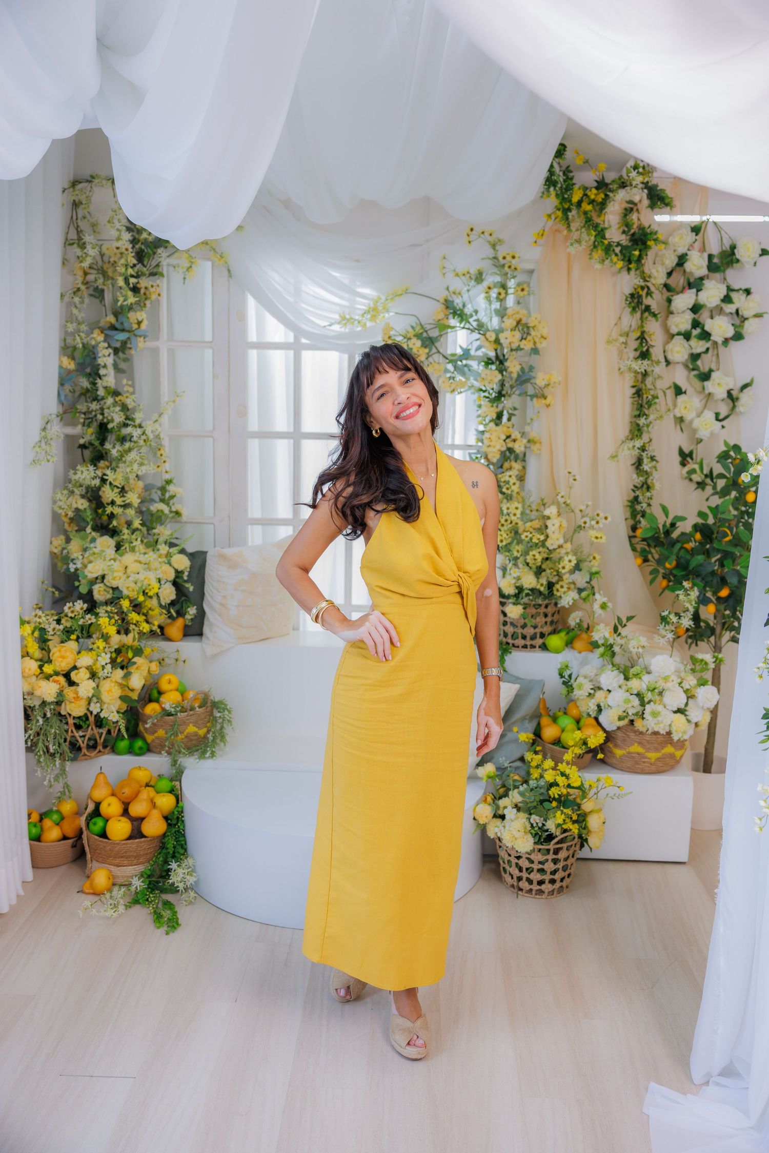 Woman in yellow maxi dress posing in floral decorated room with lemons and white flowers.