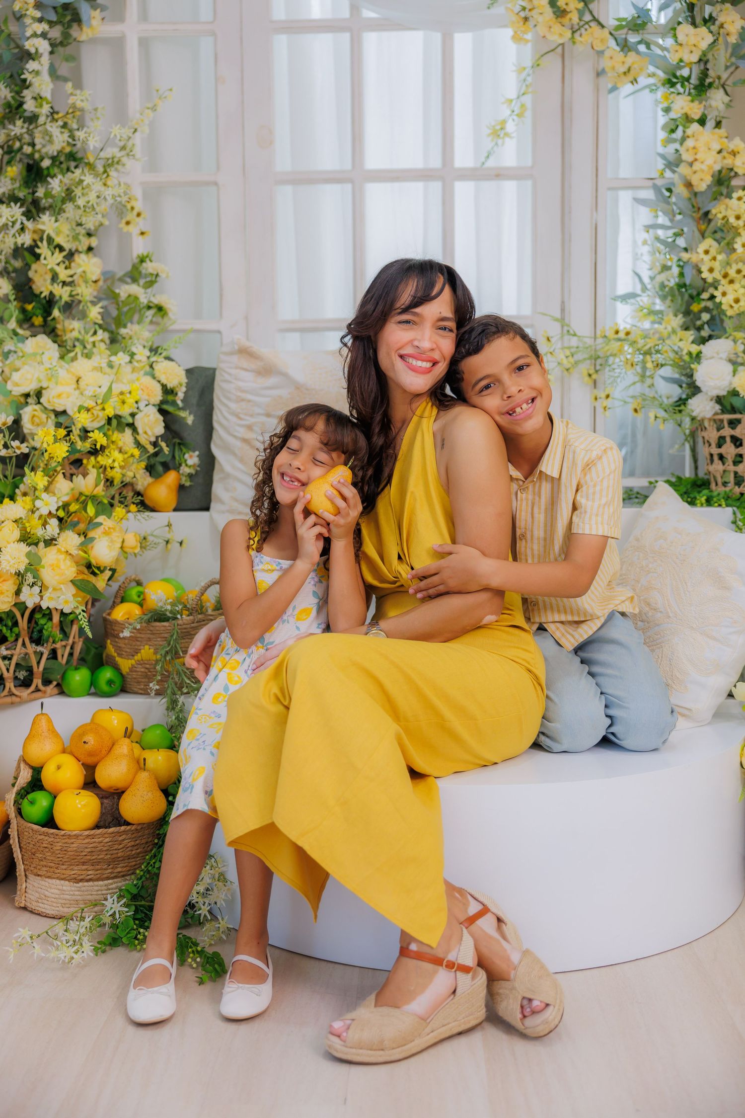 A smiling woman in yellow dress sits with two children among lemon decorations and white flowers in a bright studio.