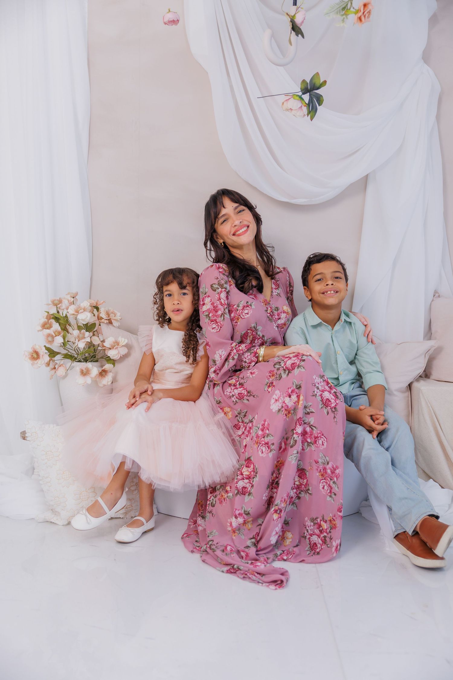A smiling woman in a floral pink dress poses with two young children in a bright, flower-decorated studio setting.
