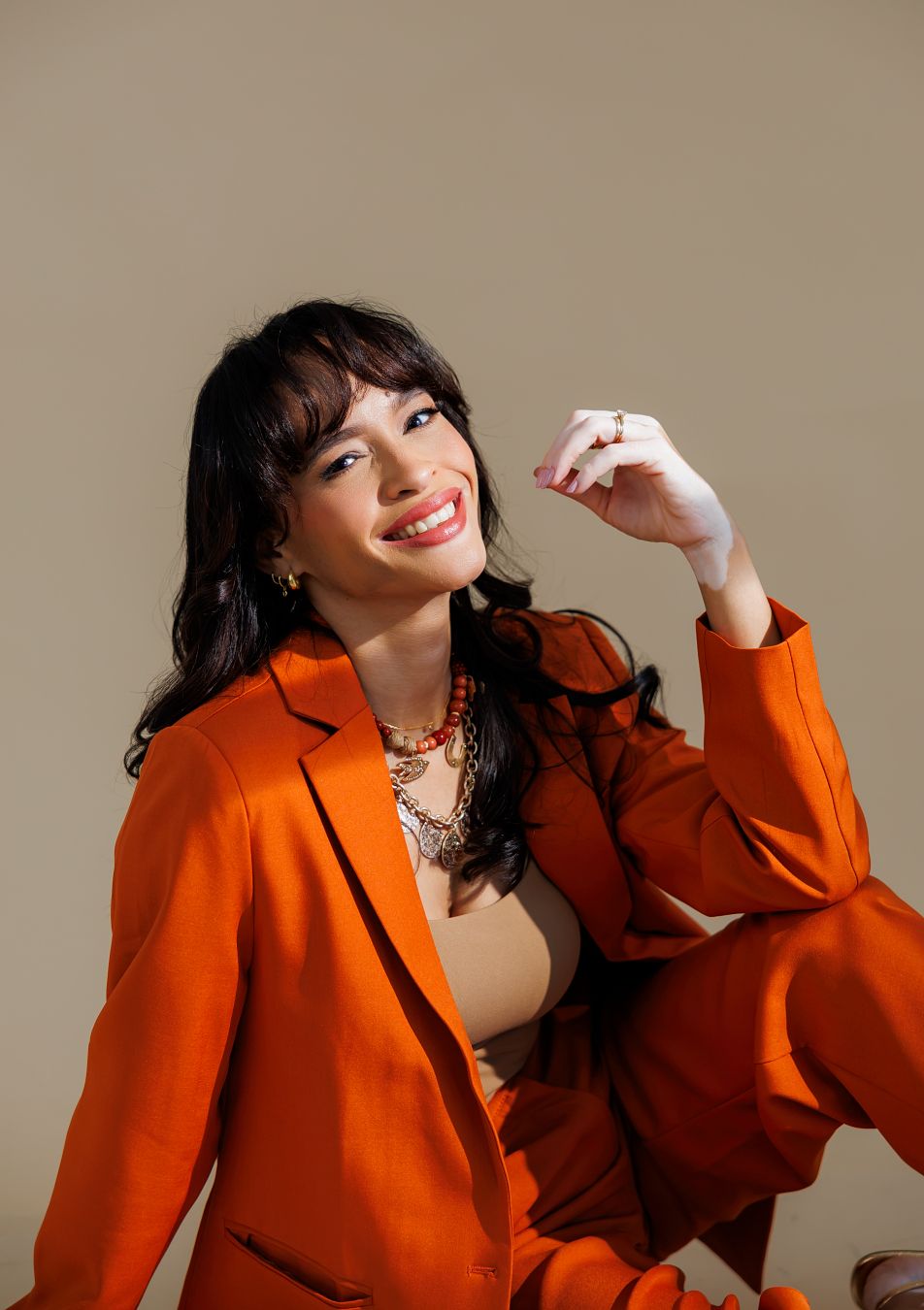 A smiling woman in an orange blazer suit poses confidently against a neutral background, wearing gold jewelry.