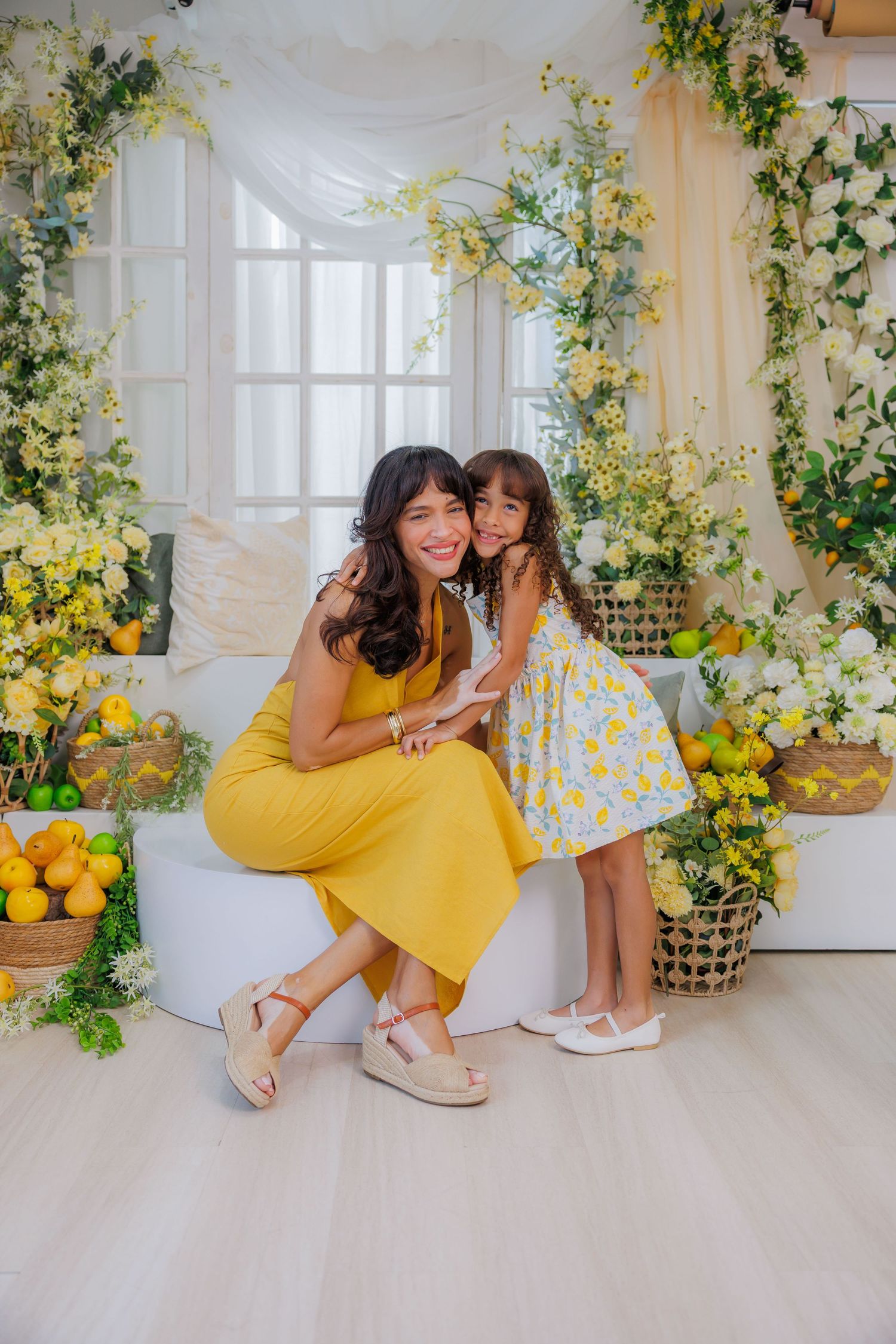 A woman in yellow dress hugs a little girl in floral dress, surrounded by yellow flowers and lemons in a bright studio.