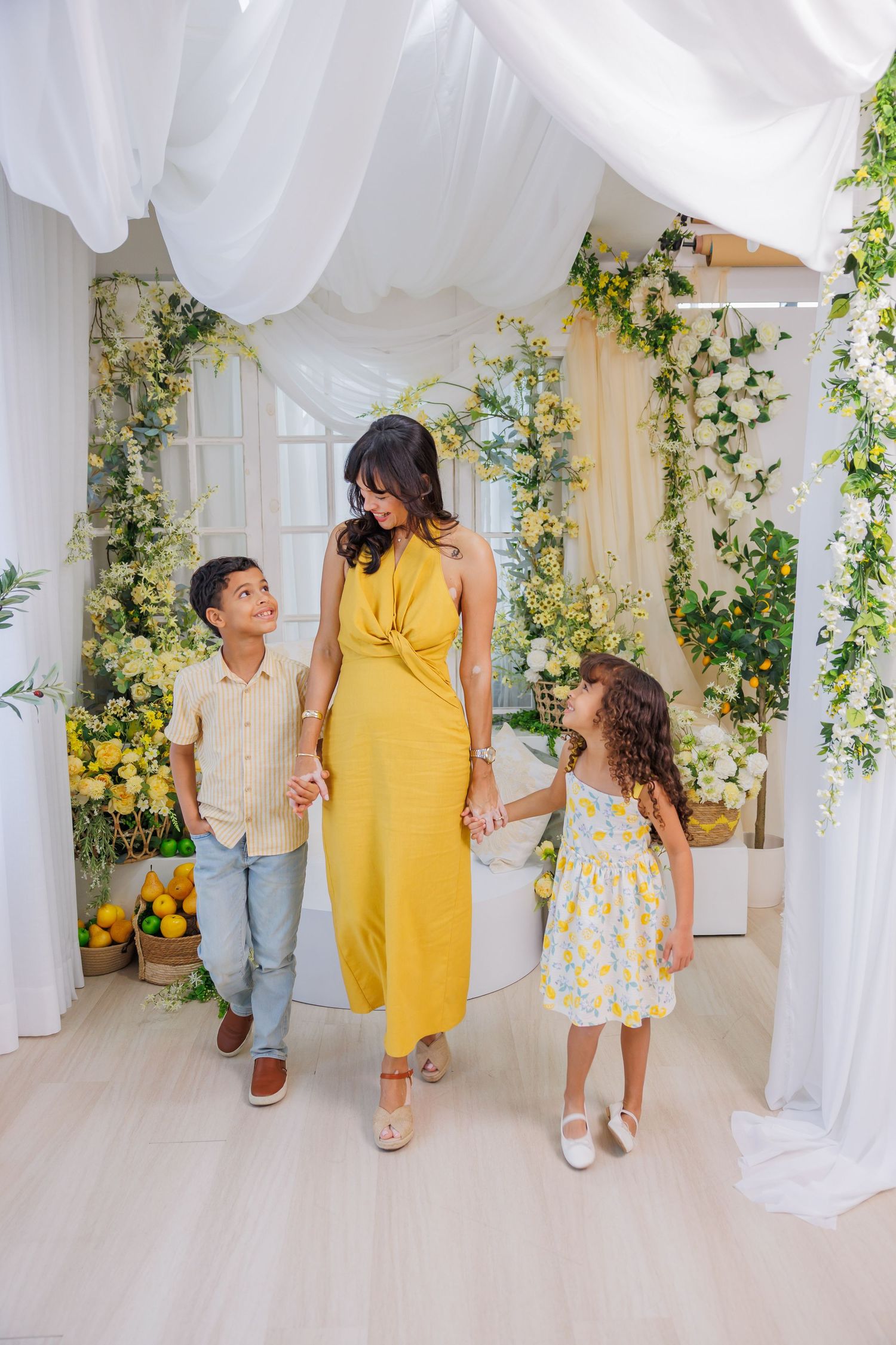 Woman in yellow dress walks with two children through floral arch with white draping and lemon decor.