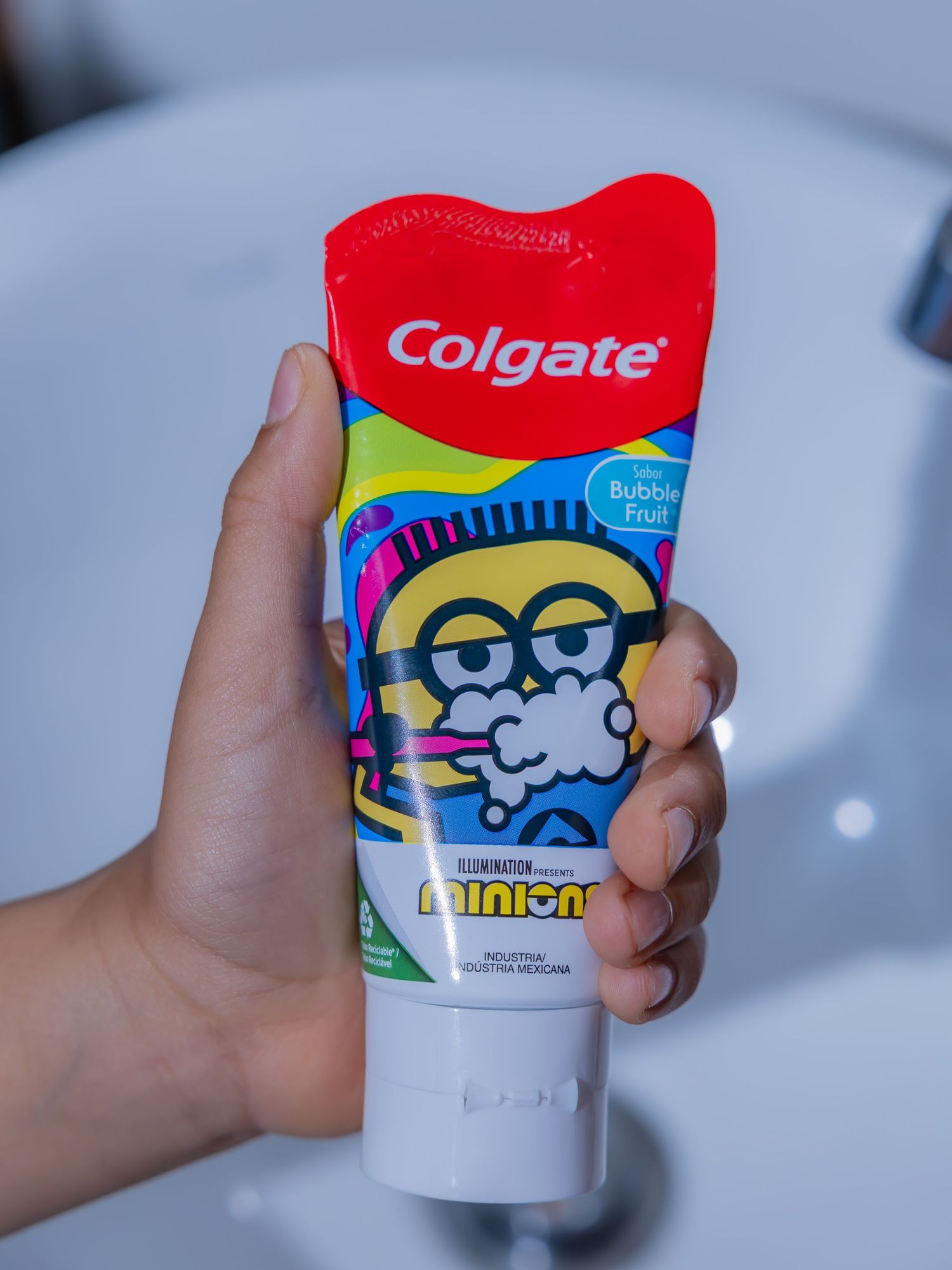 Hand holding Colgate Minions Bubble Fruit flavored kids toothpaste tube in a bathroom setting.