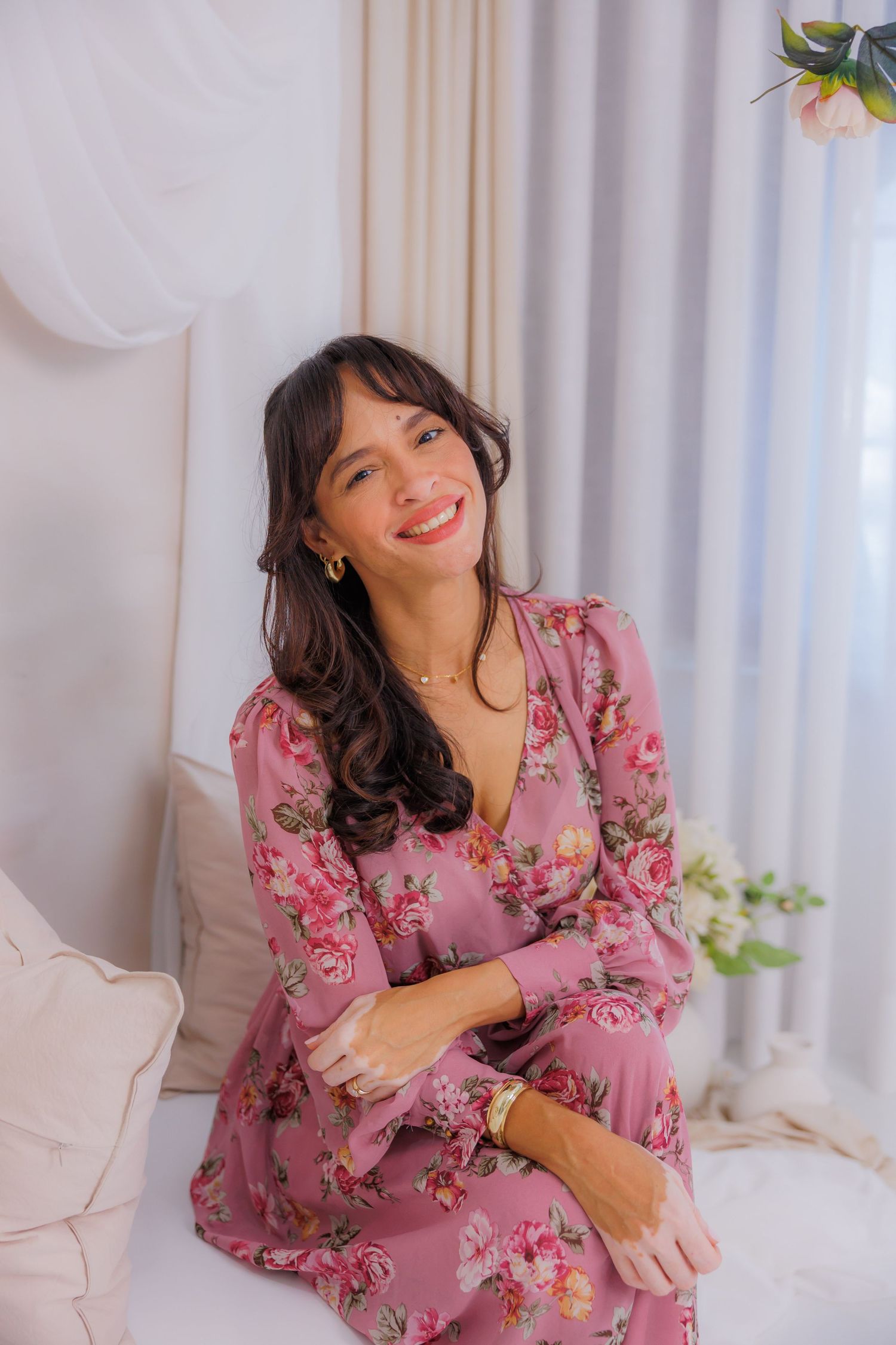 Smiling woman in pink floral long-sleeve dress sitting on a bed in a bright, elegant room with white curtains.
