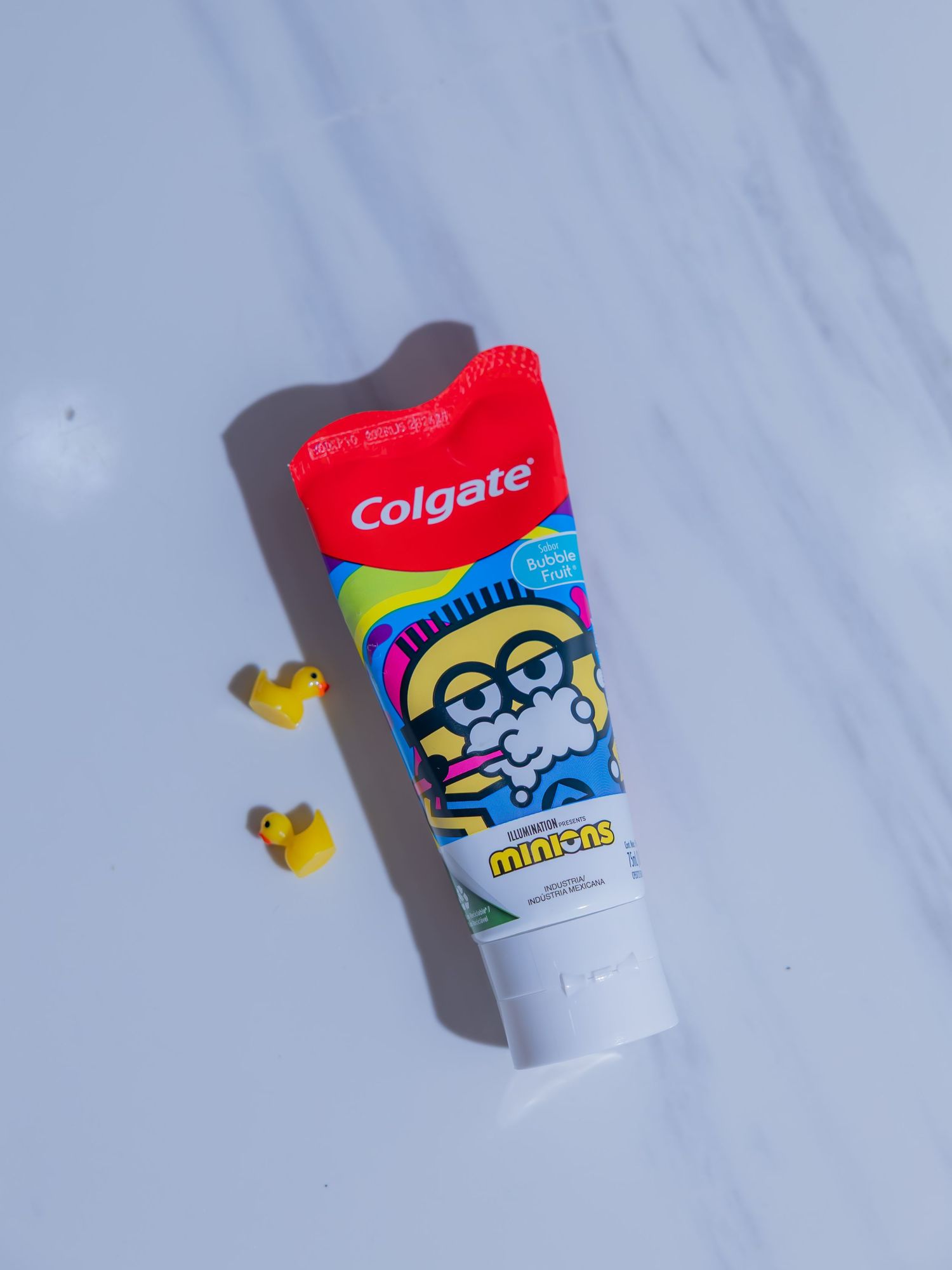 Colgate Minions kids toothpaste tube on white marble surface with two small yellow rubber duck decorations.