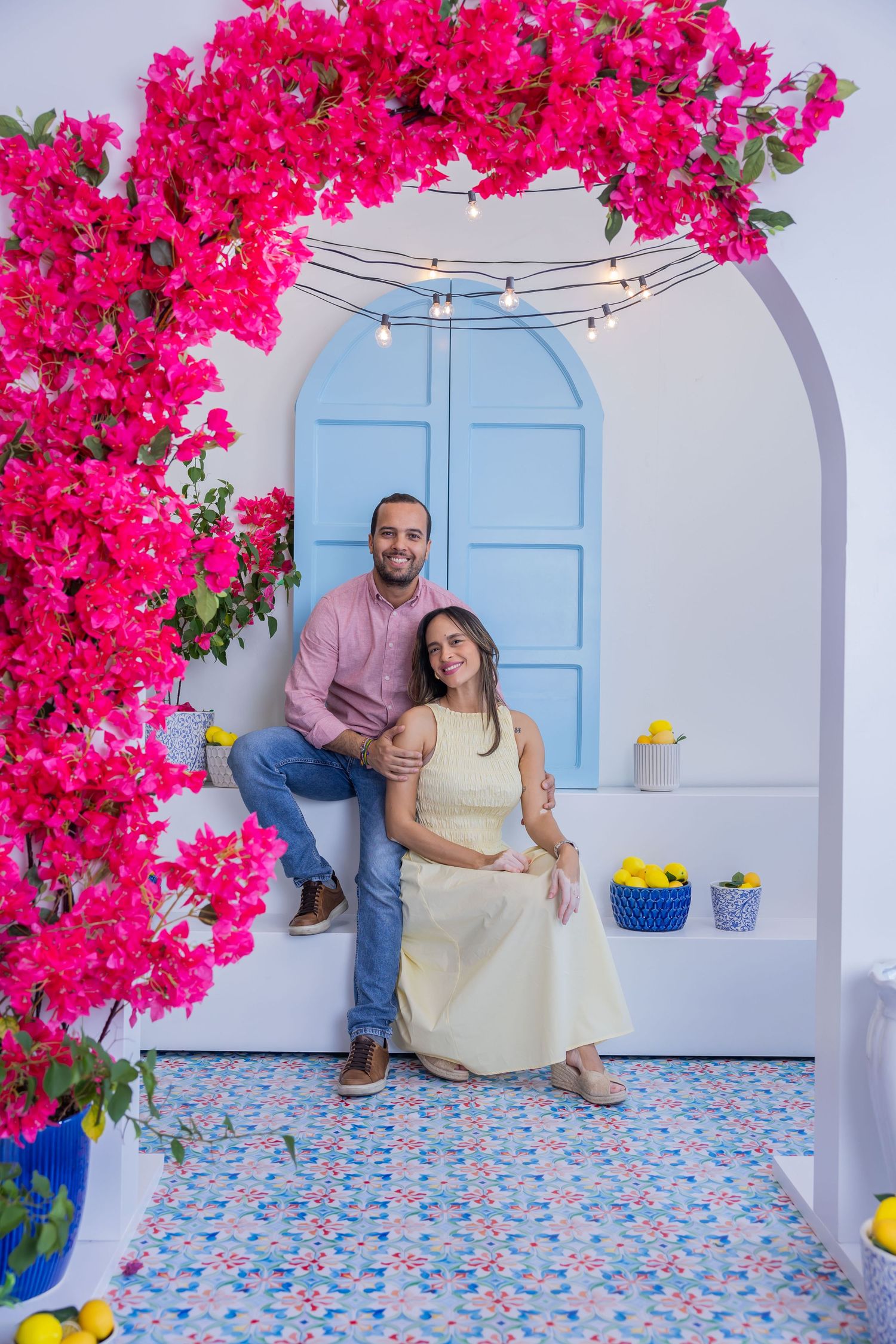 Couple posing under a pink bougainvillea arch by a blue door with string lights and lemons on a tiled patio.