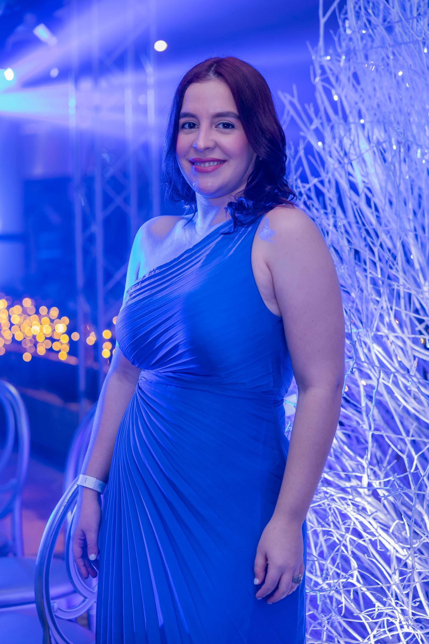 A woman in a royal blue sleeveless dress poses against a backdrop of twinkling white lights and blue ambiance.