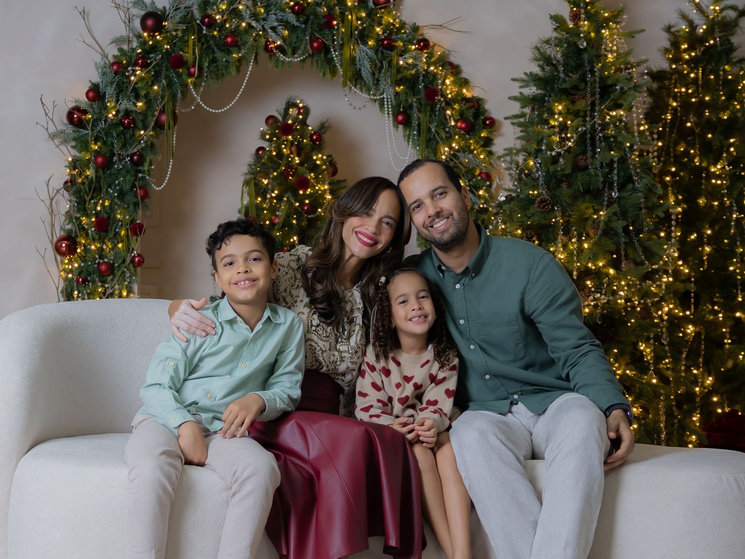 Family of four smiling together on white sofa with Christmas trees and festive wreath backdrop during holiday photo session.