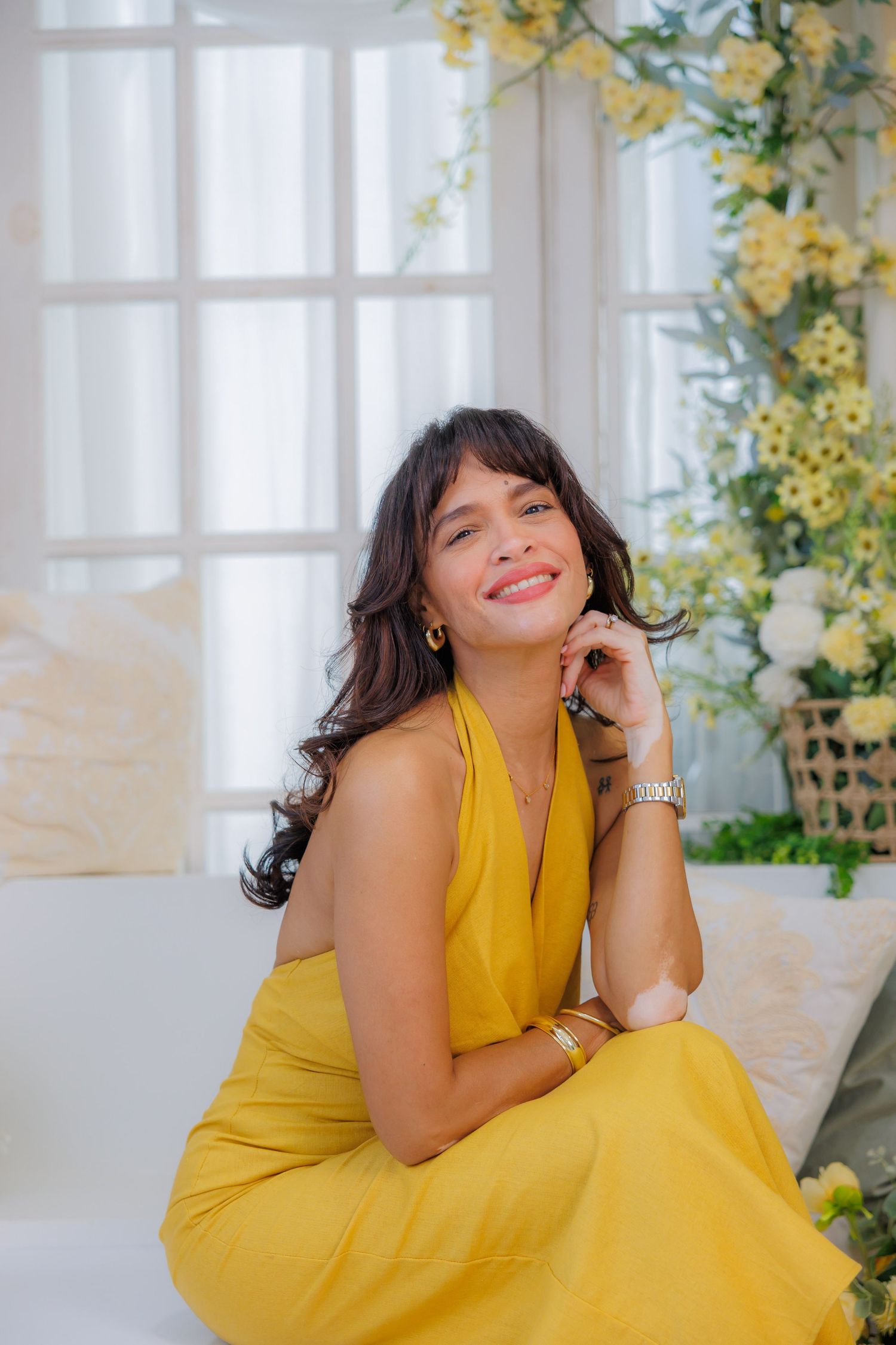 Woman in yellow halter dress sitting gracefully among white and yellow floral decorations, smiling warmly at camera.