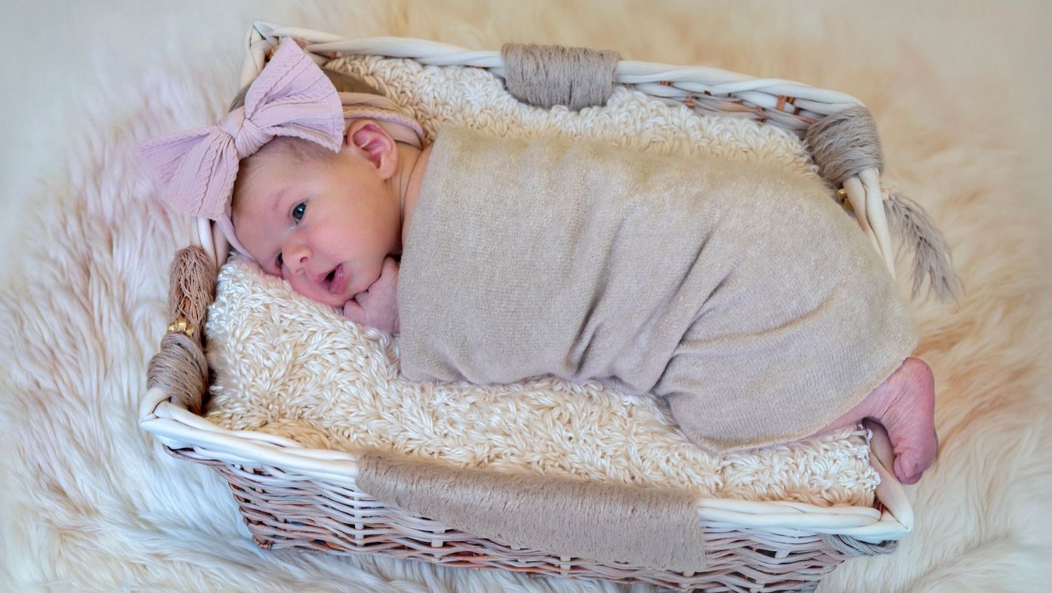 Newborn baby wrapped in soft gray swaddle with pink bow headband resting in white wicker basket on fluffy rug.