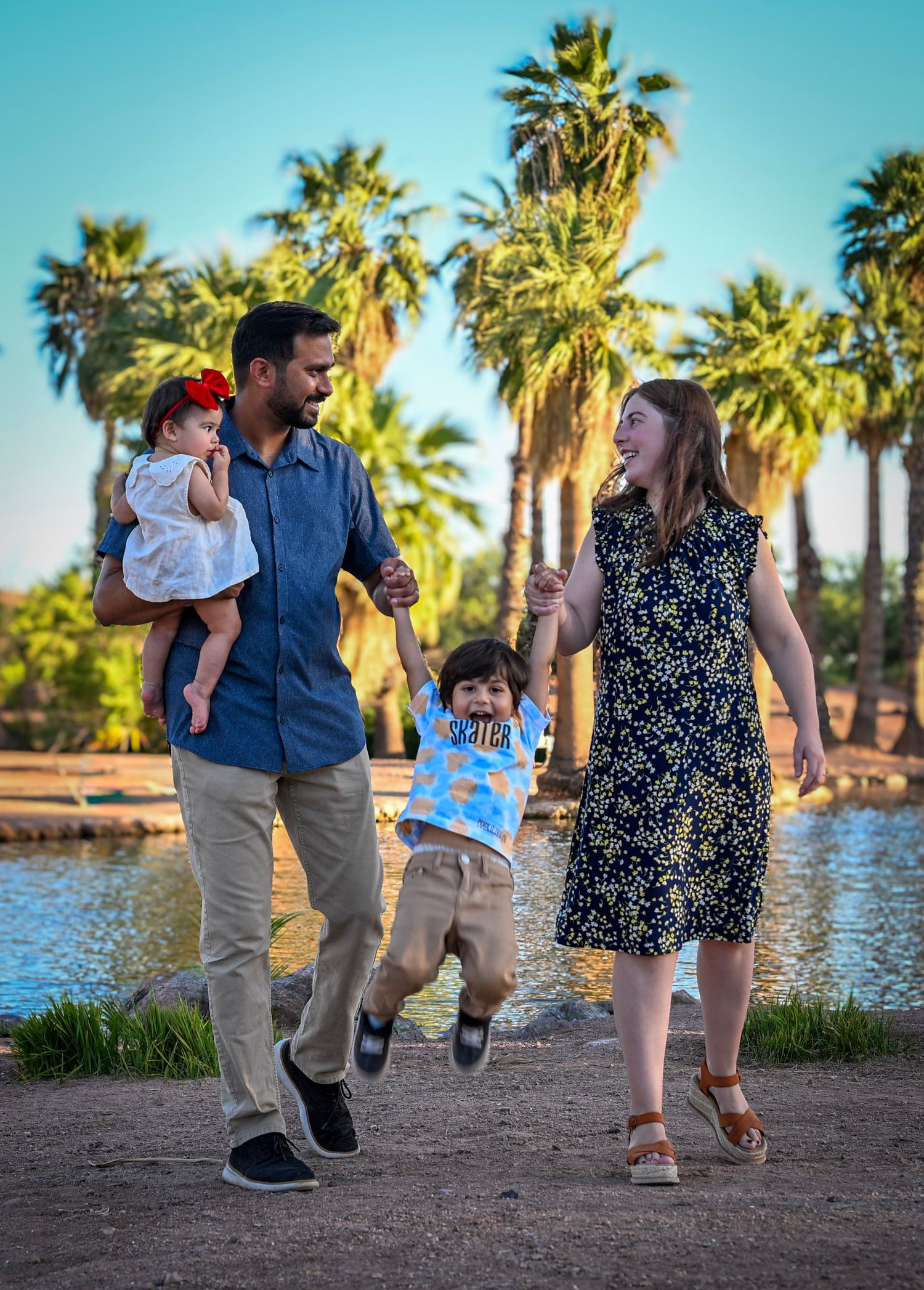 Happy family with two young children swinging toddler between parents near palm tree lined lake on sunny day.