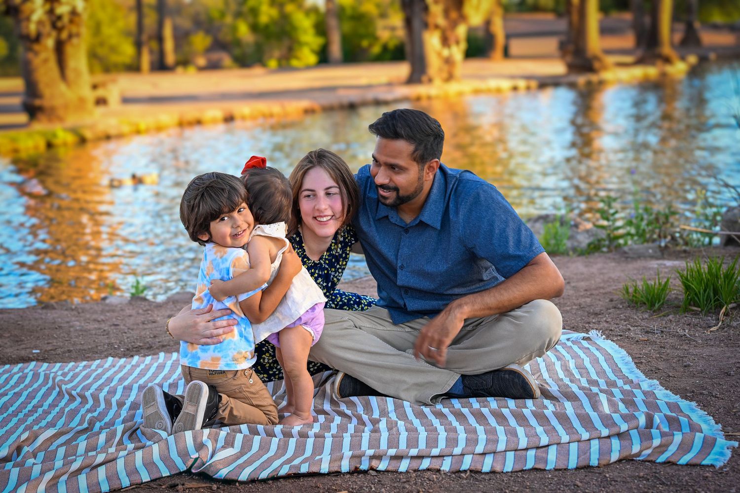 Happy family of three sitting on a blanket by a pond in a sunny park, smiling and embracing together.