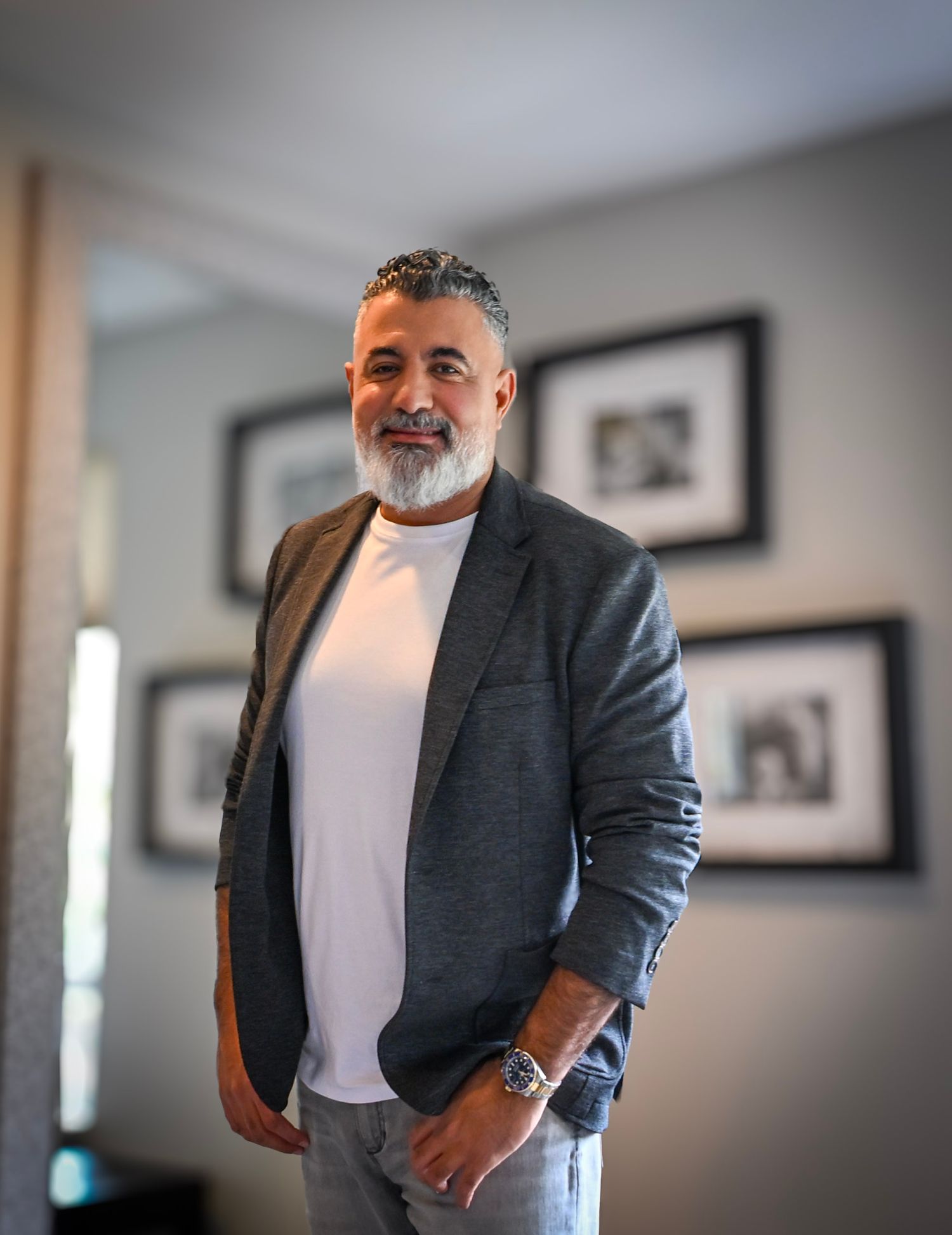 A person in a gray blazer and white t-shirt poses against a blurred interior background with framed artwork.