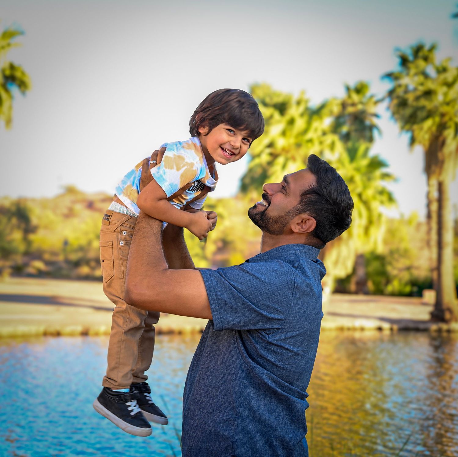 A man joyfully lifts a laughing toddler boy outdoors near a pond with palm trees in the background.