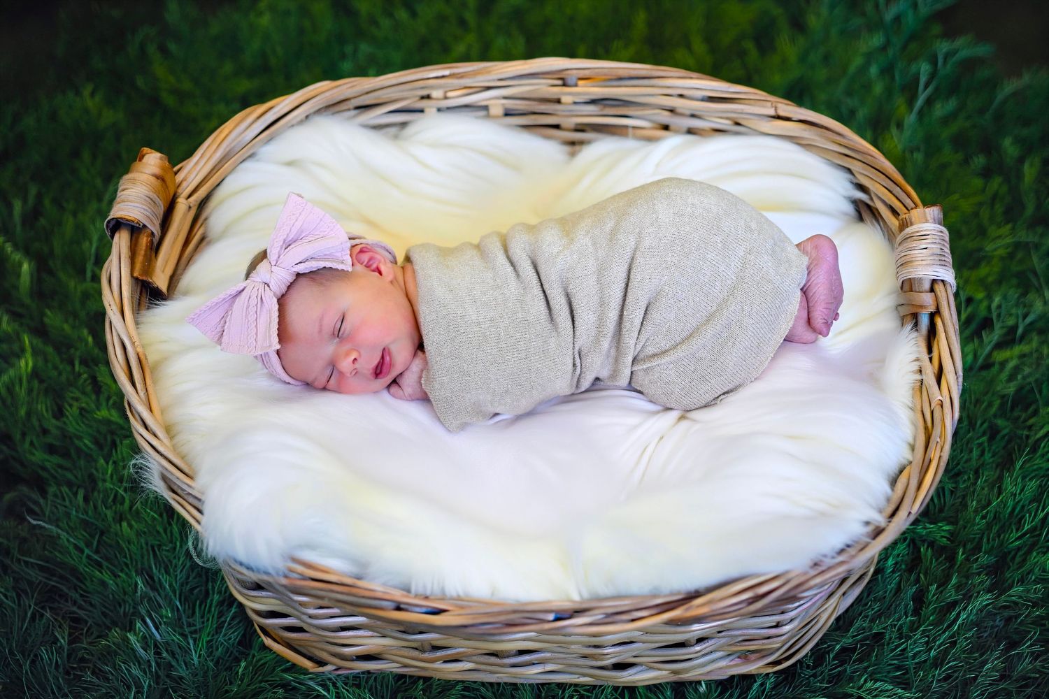 Newborn baby swaddled in gray wrap with pink bow headband sleeping in wicker basket on white fur outdoors.