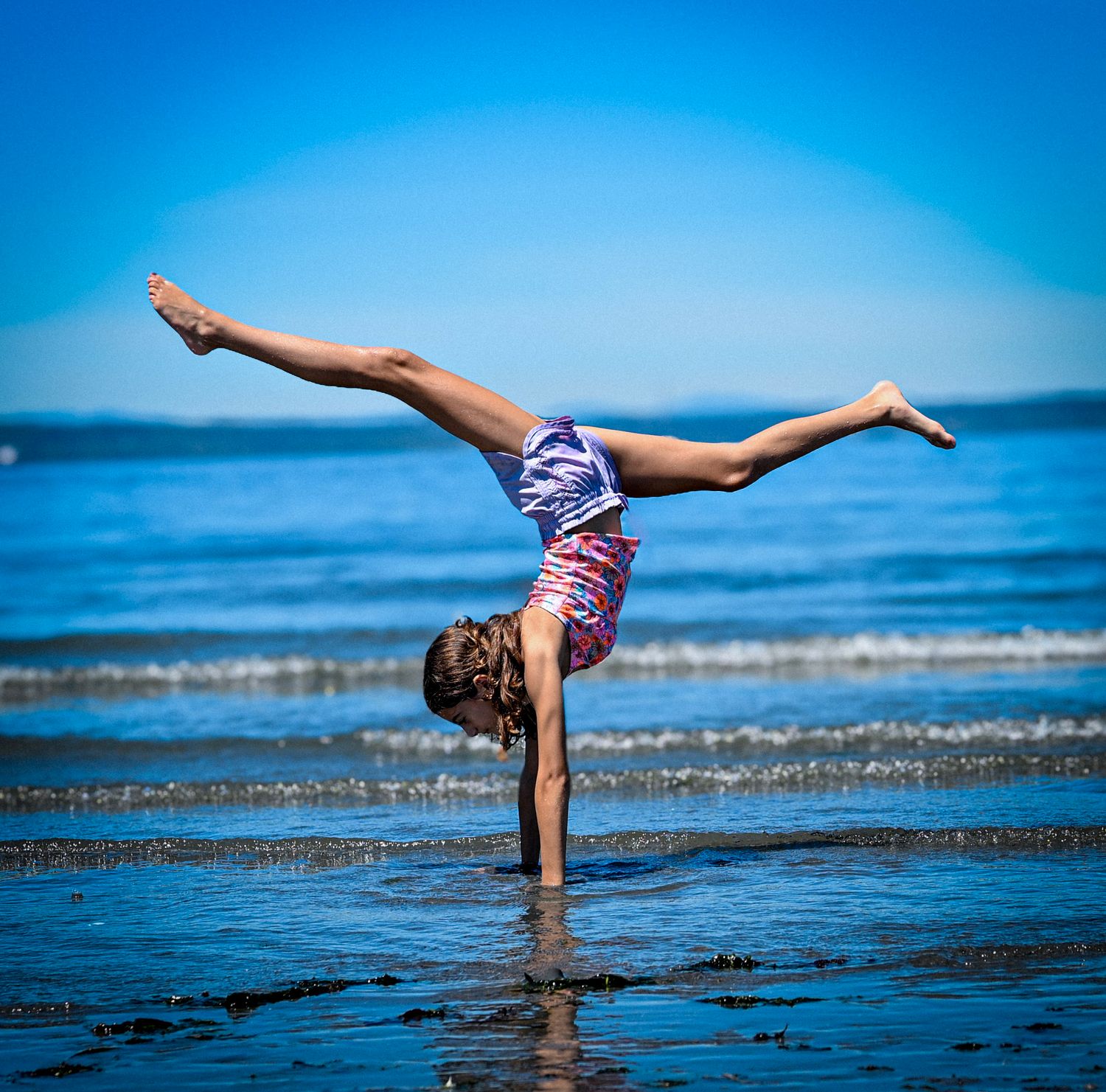 A gymnast performs a one-handed handstand split on a wet beach with blue ocean waves in the background.